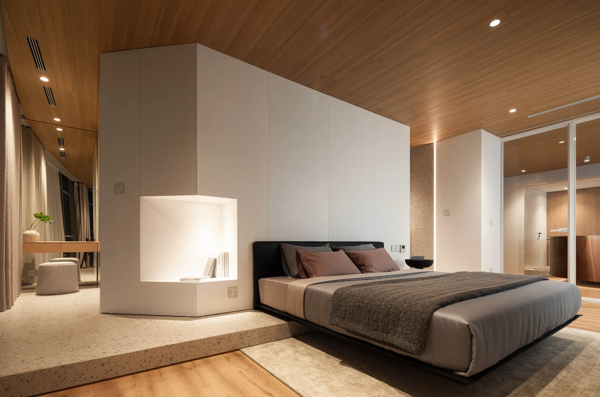 A low-profile bed sits against a large white partition wall with an integrated lighted niche, beneath a warm wood-planked ceiling.