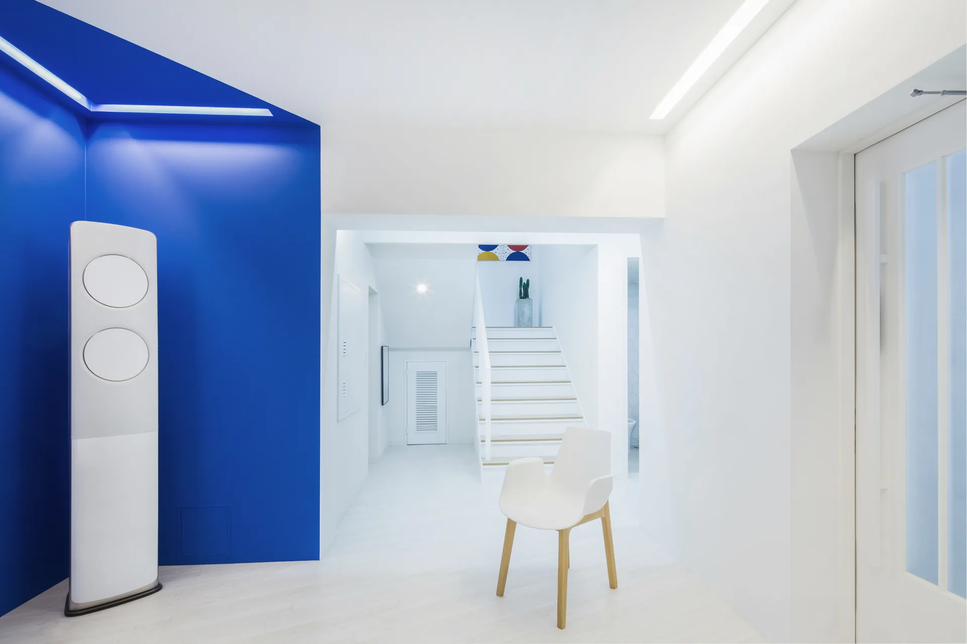 A minimalist white entryway featuring a bold blue accent wall, a white armchair, and a staircase in the background.