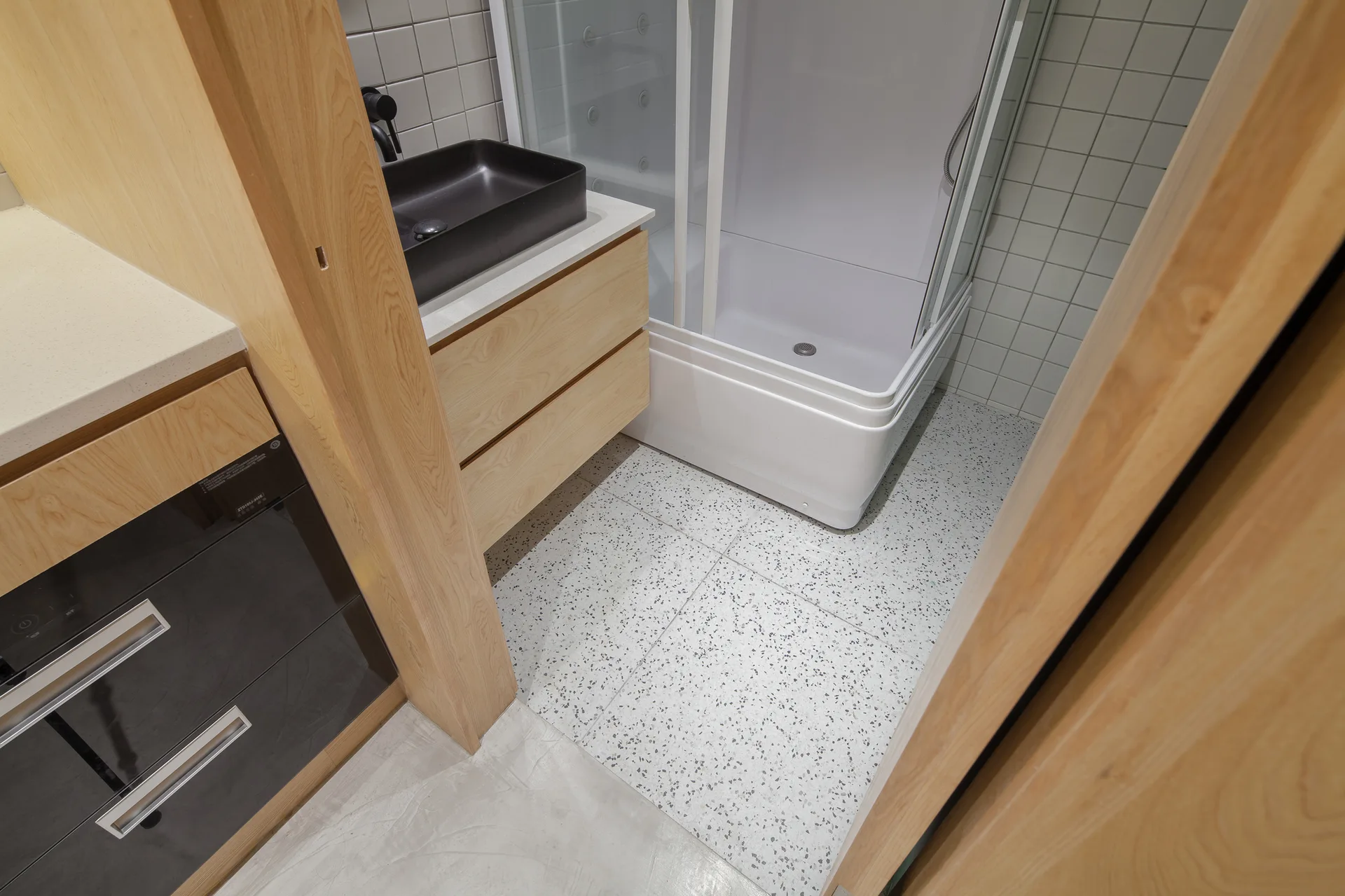 A modern bathroom with a light wood vanity, black vessel sink, white square tiled walls, a glass shower stall, and terrazzo floor tile.