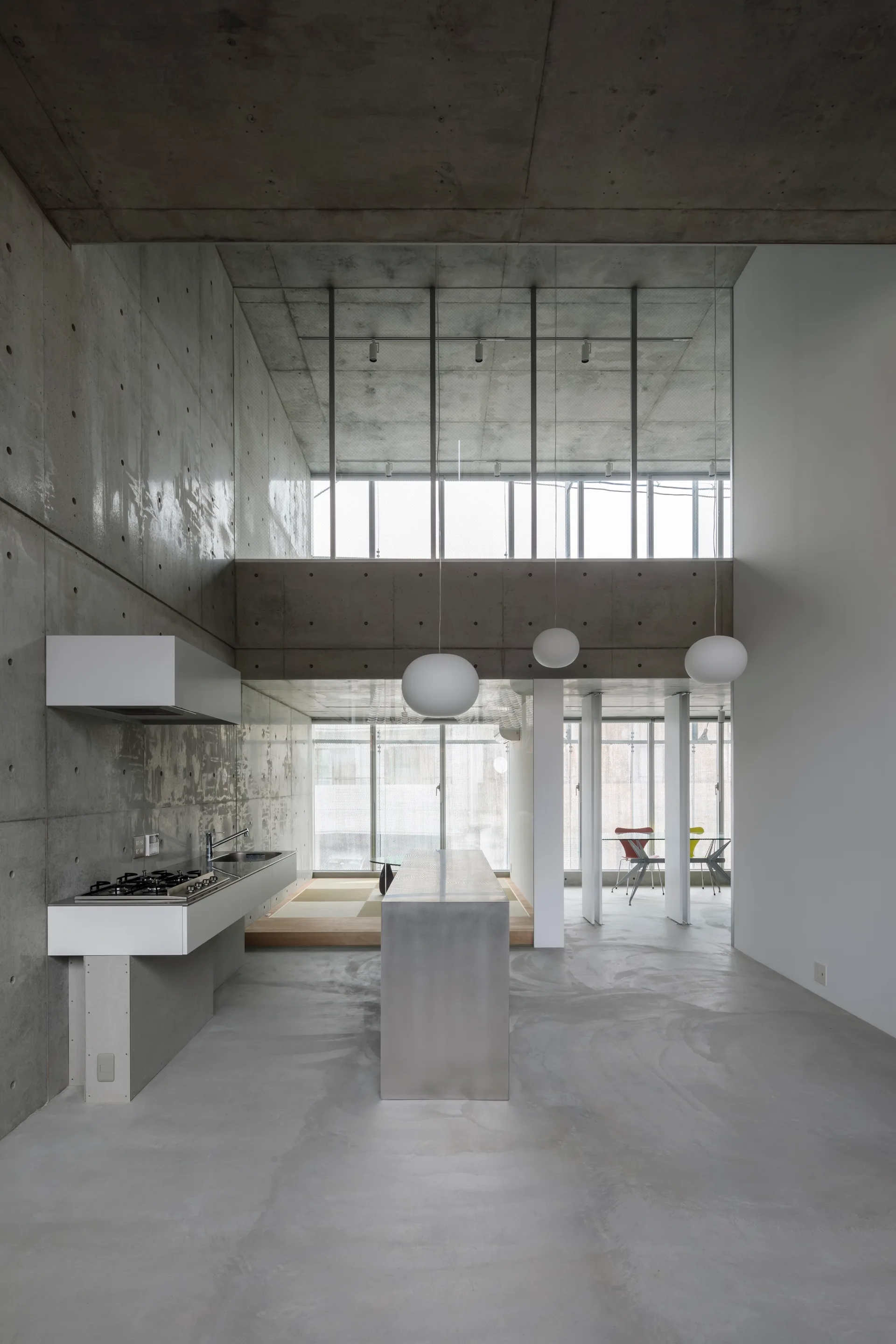 A minimalist industrial kitchen featuring raw concrete walls, a metallic stainless steel island, and globe pendant lights.