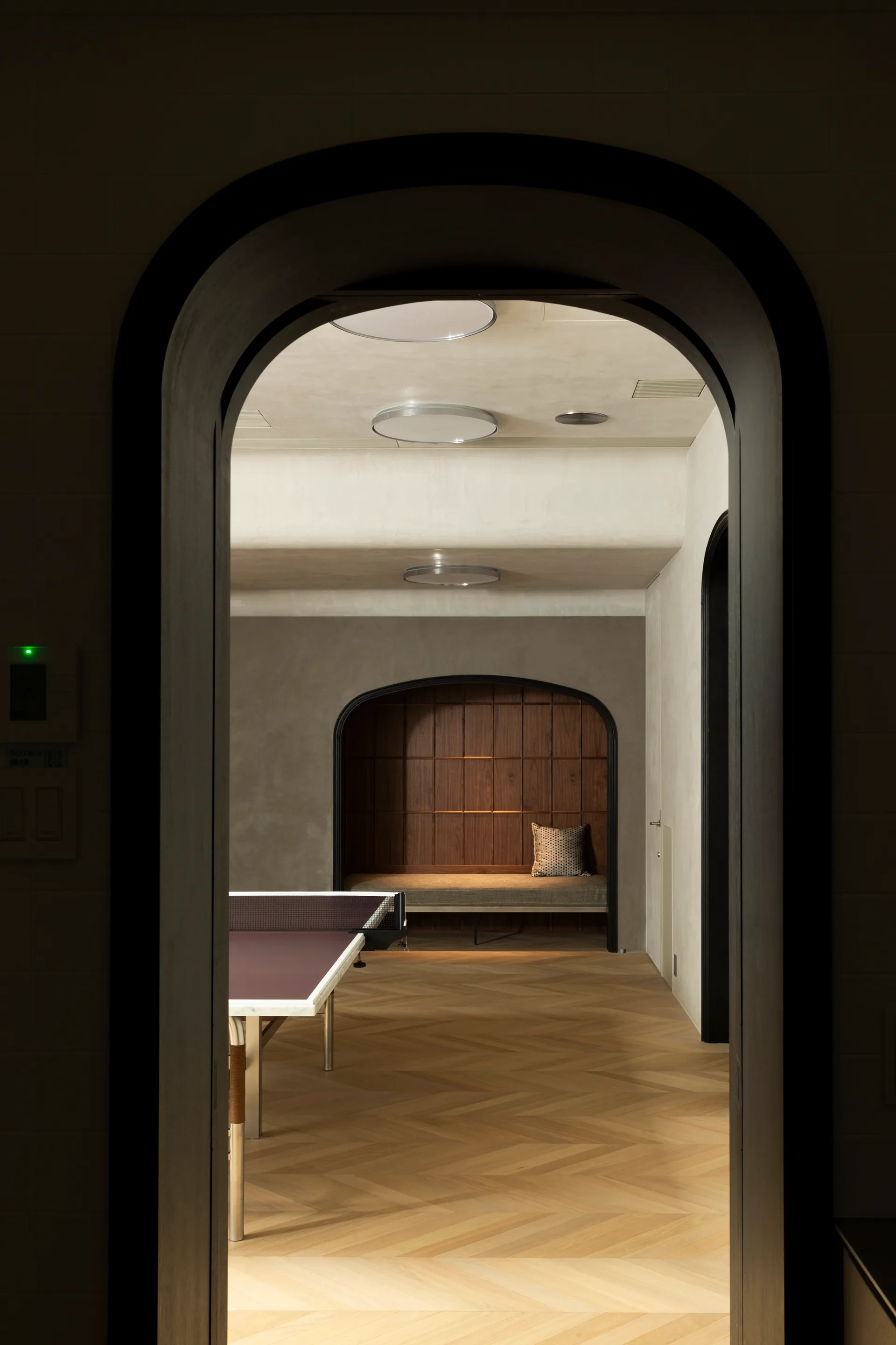 A view through a dark arched doorway showing a game room with herringbone wood floors, a ping pong table, and a recessed wooden seating alcove.