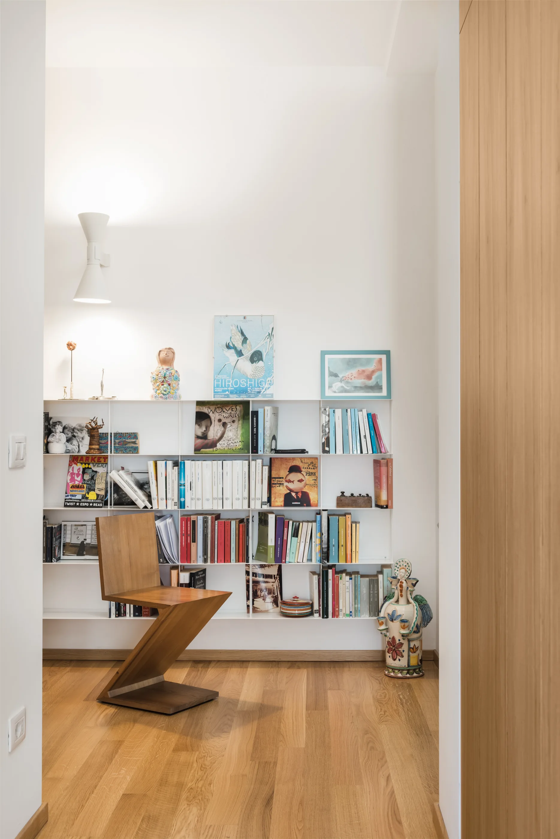 A wooden zig-zag chair sits in front of a white grid bookshelf filled with books and colorful artwork on a hardwood floor.