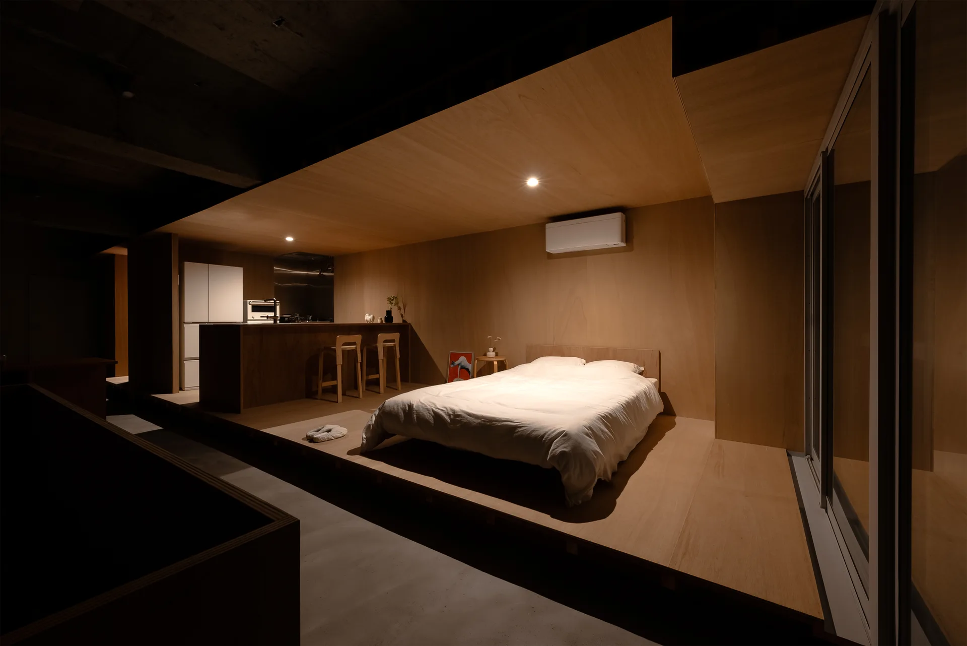 A minimalist bedroom with a bed on a raised plywood platform next to a kitchen area with wood-paneled walls and ceiling.