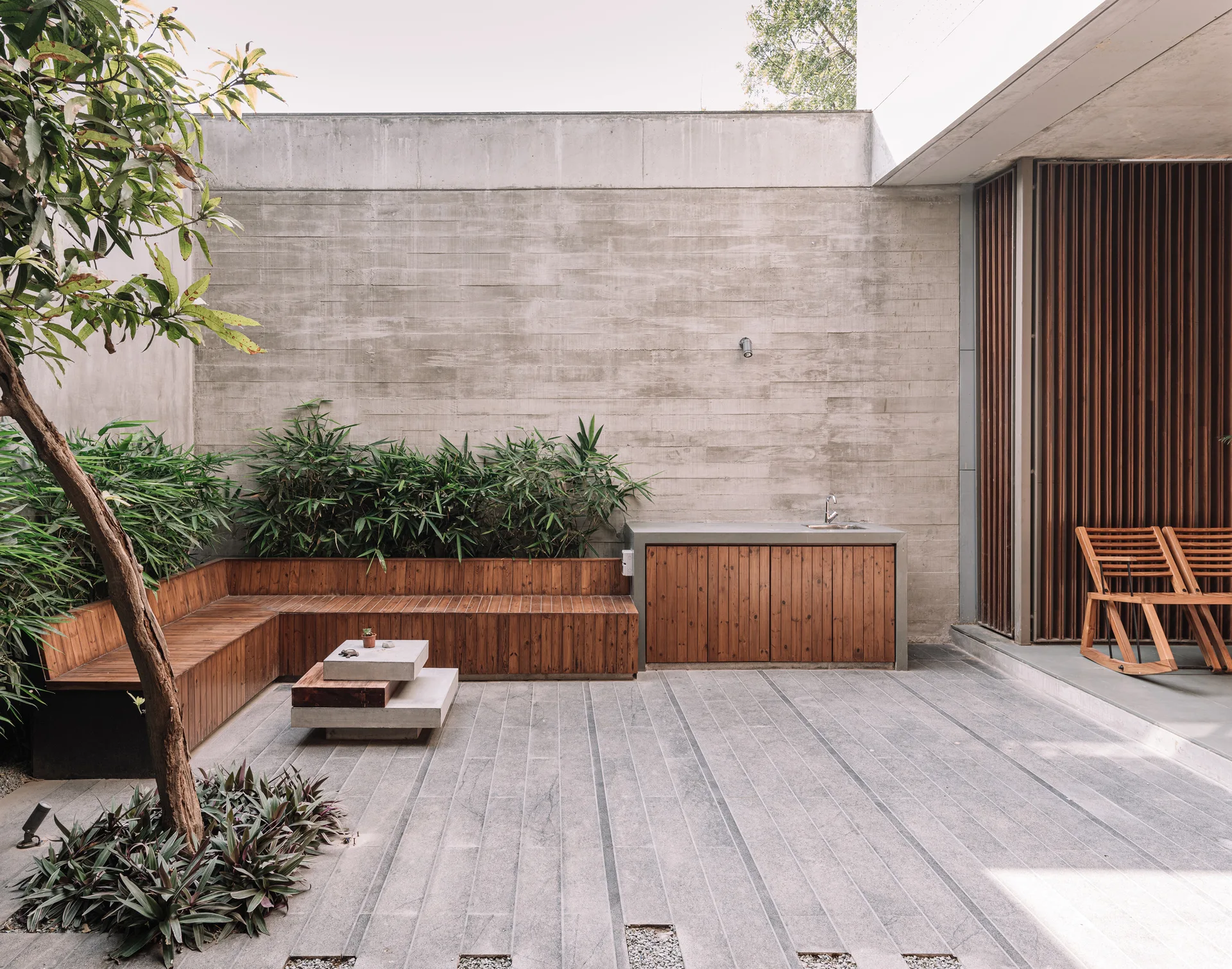 An outdoor courtyard patio features a board-formed concrete wall, an L-shaped wood bench, a concrete sink console, and stone slab flooring.