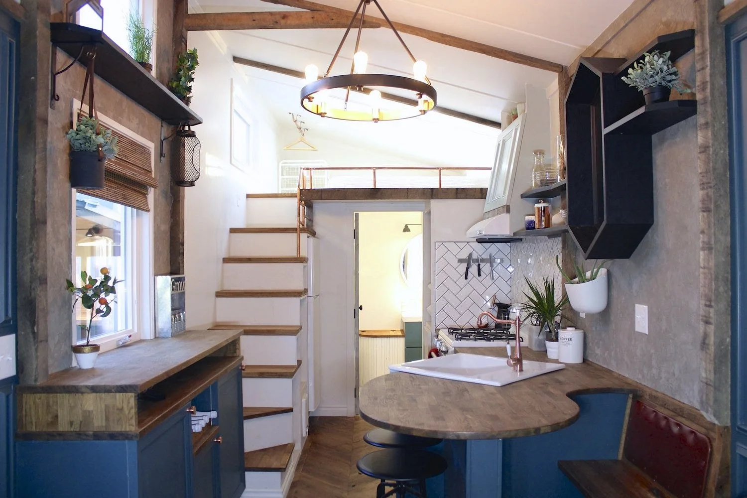 A compact tiny home kitchen with blue cabinets, wooden countertops, a white herringbone tile backsplash, and wooden stairs leading to a loft.