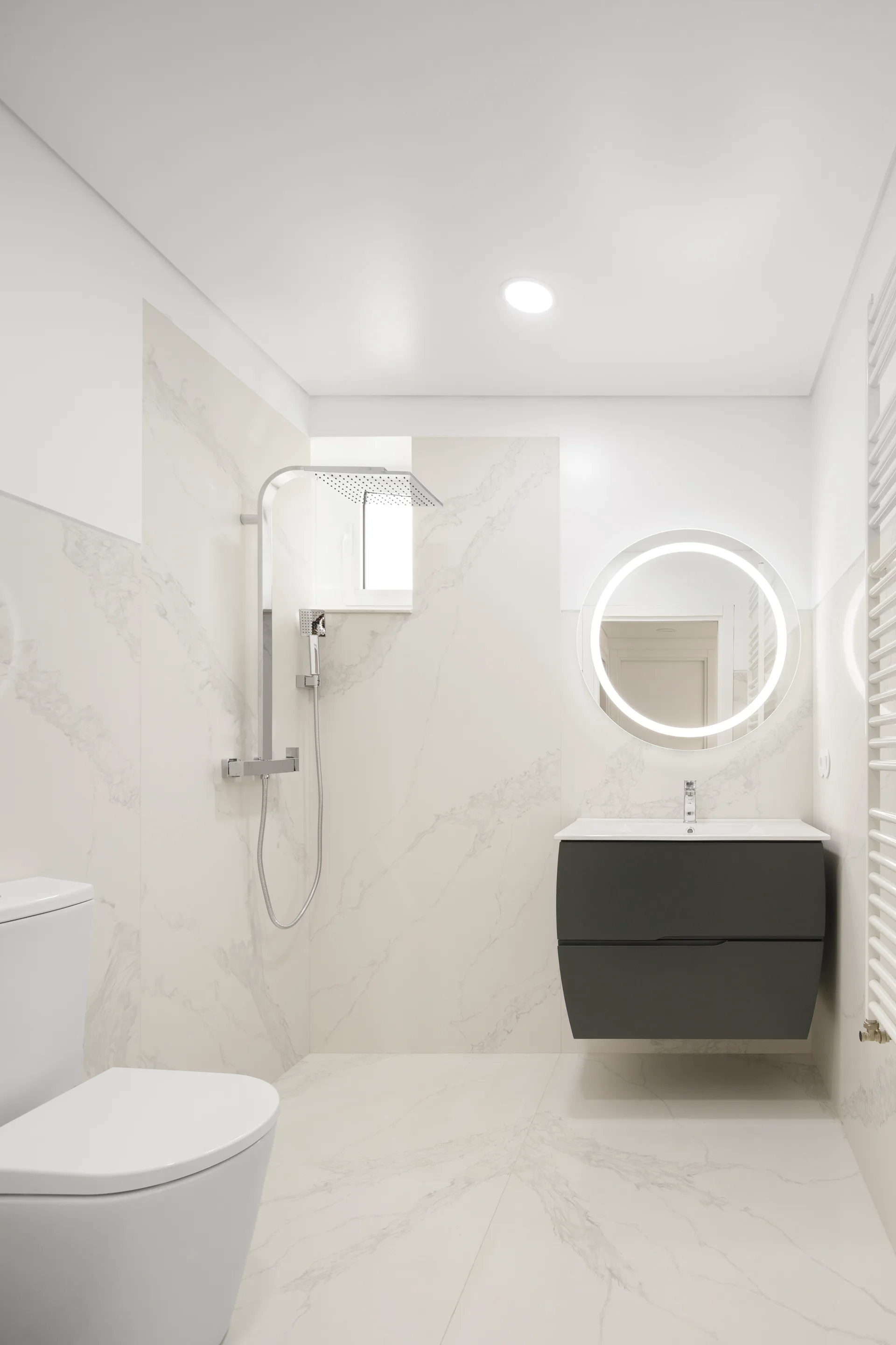 A minimalist bathroom features veined marble-look wall tiles, a round backlit LED mirror, a dark grey floating vanity, and a chrome rainfall shower.