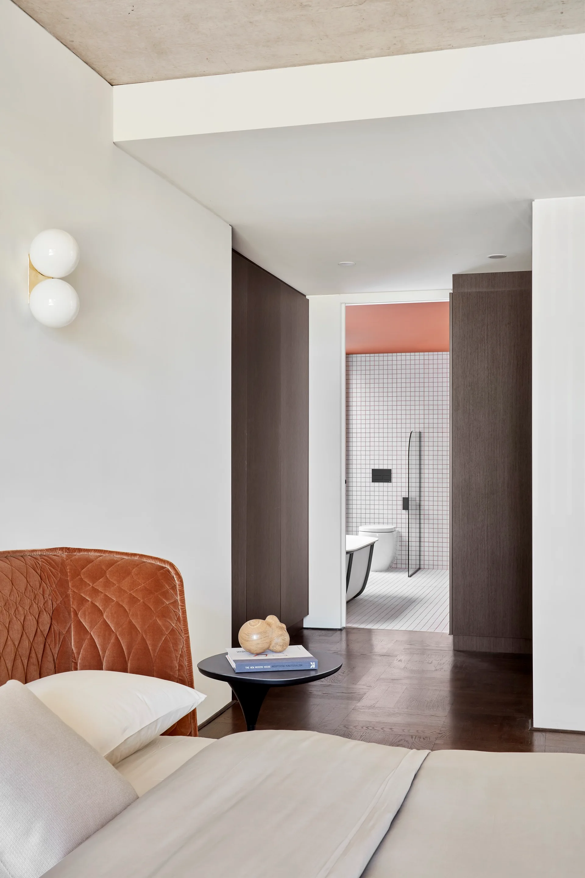 A modern bedroom with a quilted orange headboard and dark wood floors transitions into a bathroom with white grid tiles.