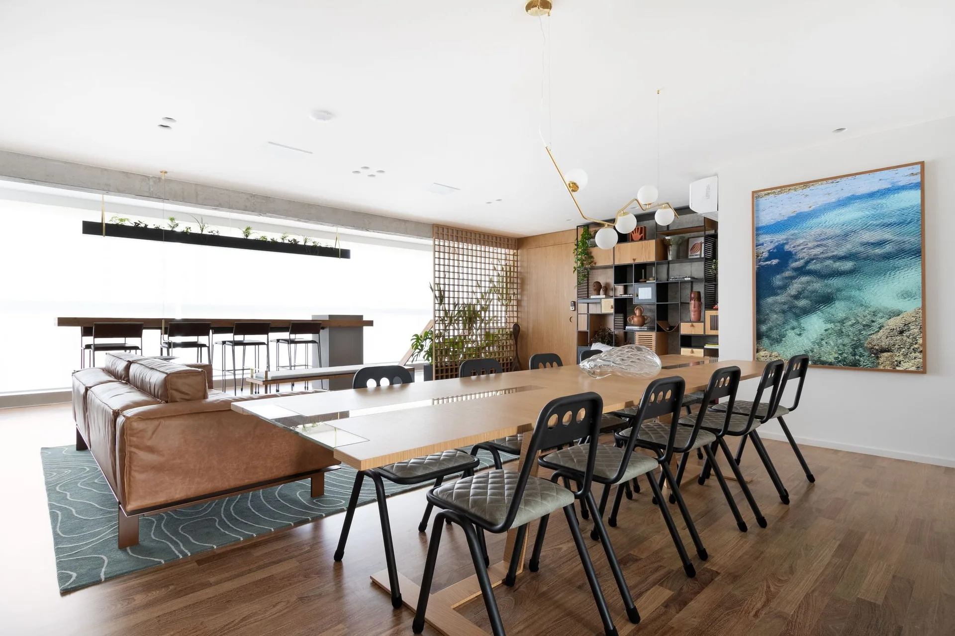 An open dining area features a long wood table with black metal chairs, a leather sofa, a wooden grid room divider, and a concrete ceiling beam.