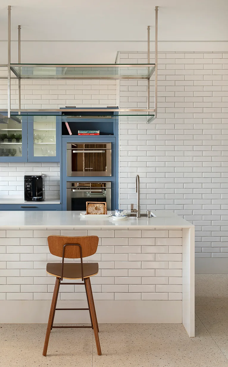 A modern kitchen featuring a white subway tiled island with a wooden stool, suspended glass shelving, and blue built-in cabinets.