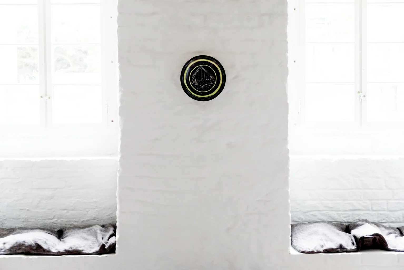 Two windows with built-in niches holding gray cushions are separated by a white painted brick wall decorated with a circular plate.