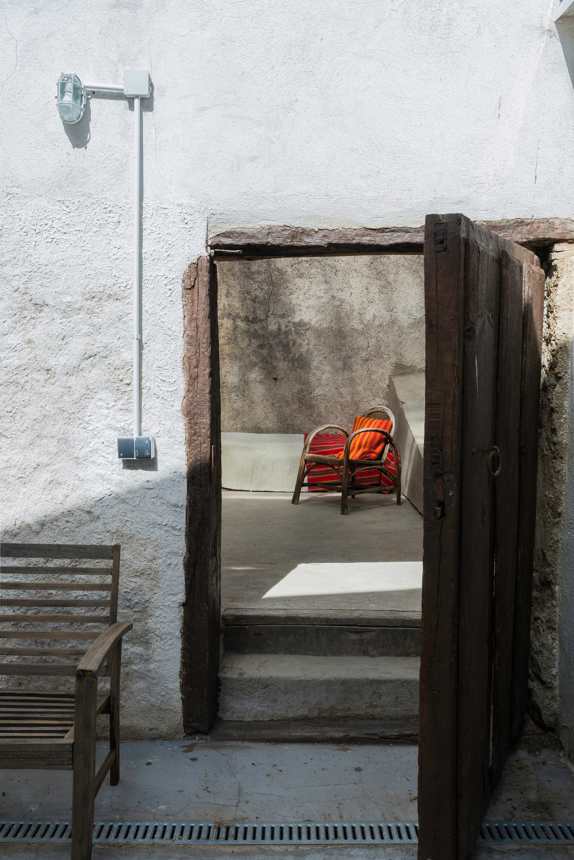 A weathered wooden doorway opens to a sunlit concrete courtyard featuring a rattan armchair with a striped orange cushion.