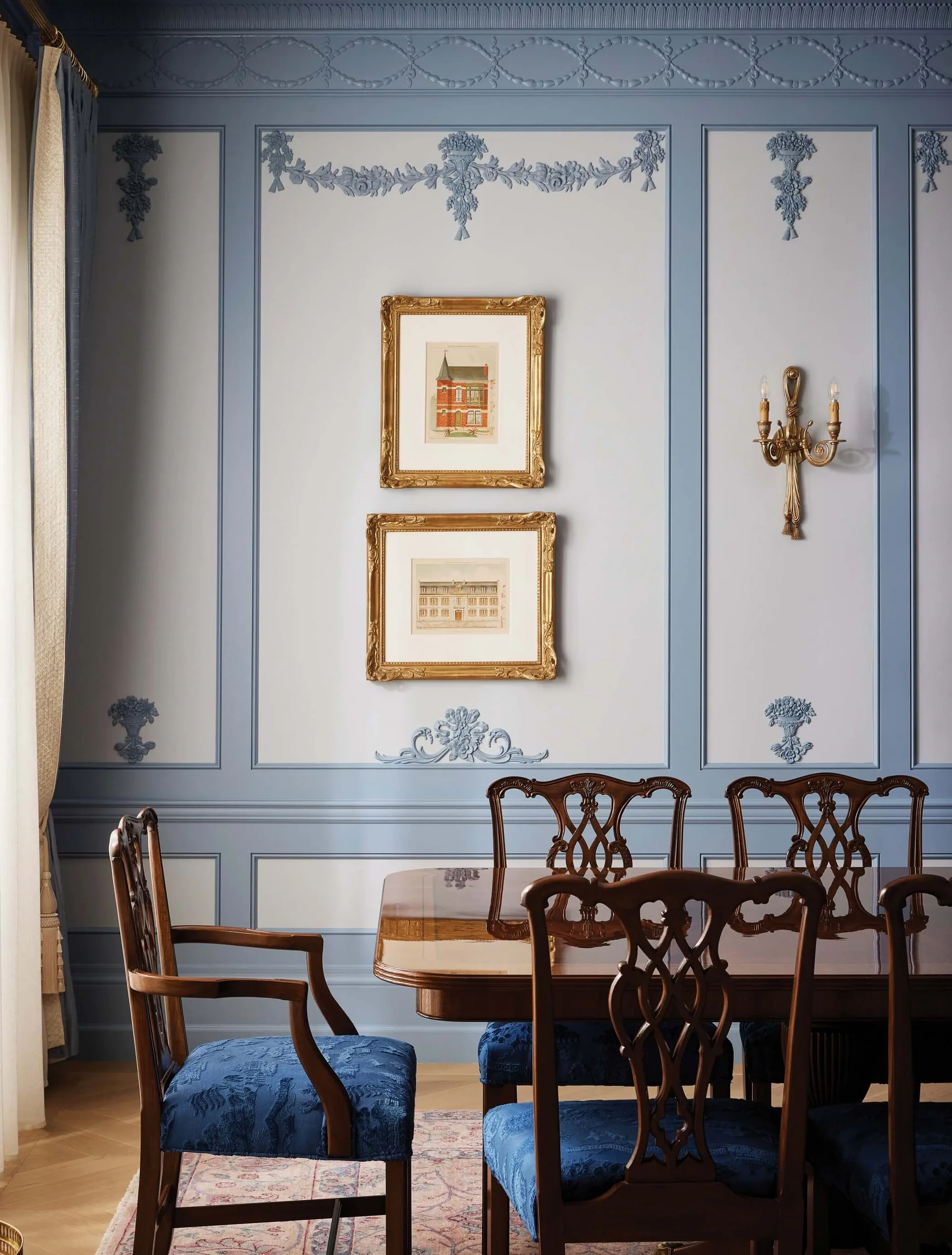 A formal dining room features a polished wood table, chairs with blue upholstery, and two-tone blue wall paneling with ornate molding and framed art.