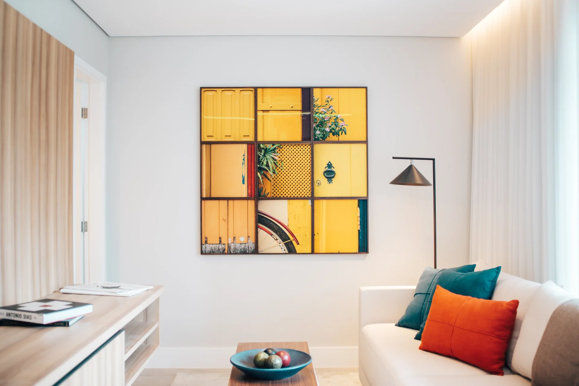 A modern living room features a white sofa with orange and teal pillows, a wooden console, and a large nine-panel yellow art piece.