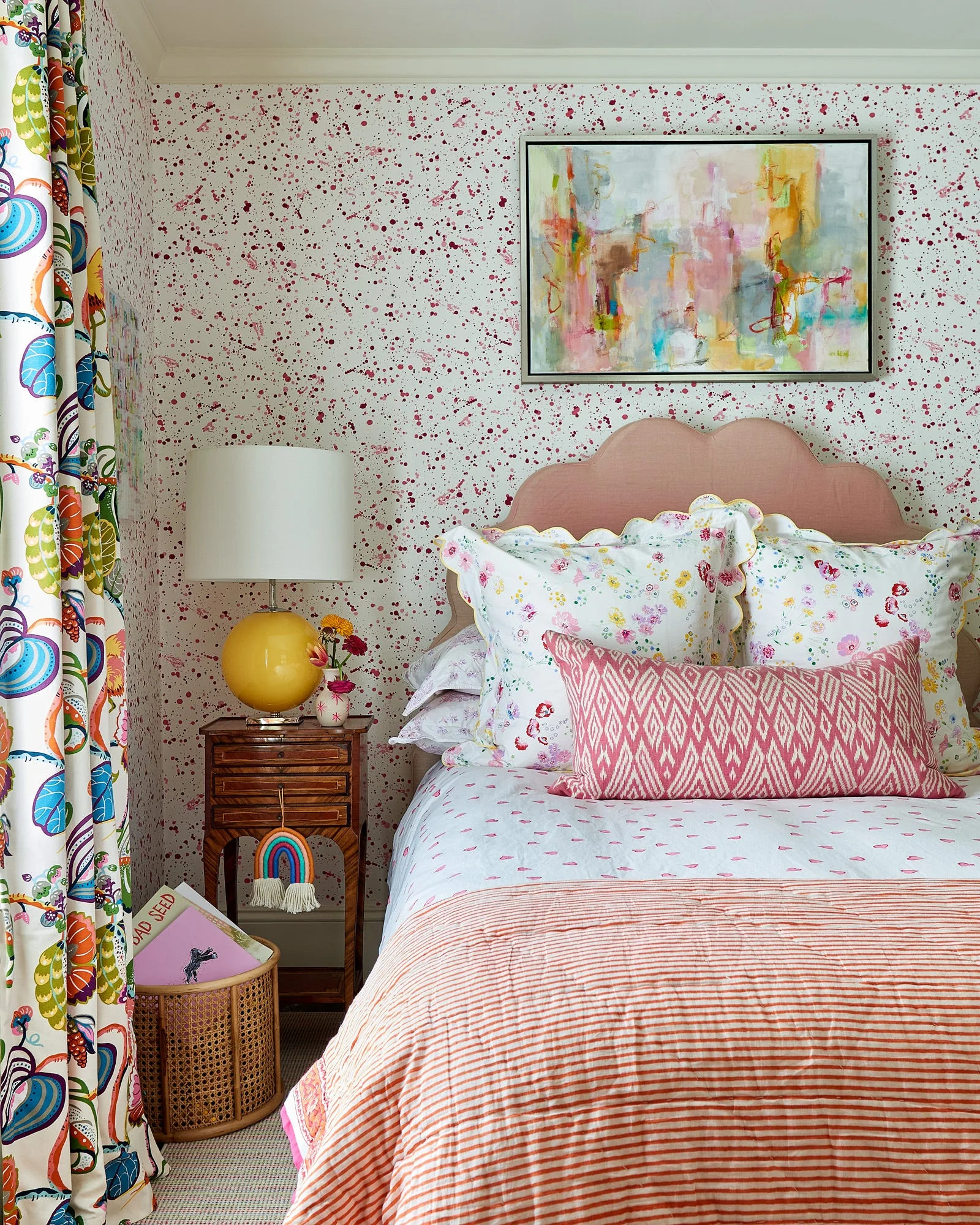 A bedroom features a scalloped pink headboard, floral pillows, and striped bedding set against pink splattered wallpaper.
