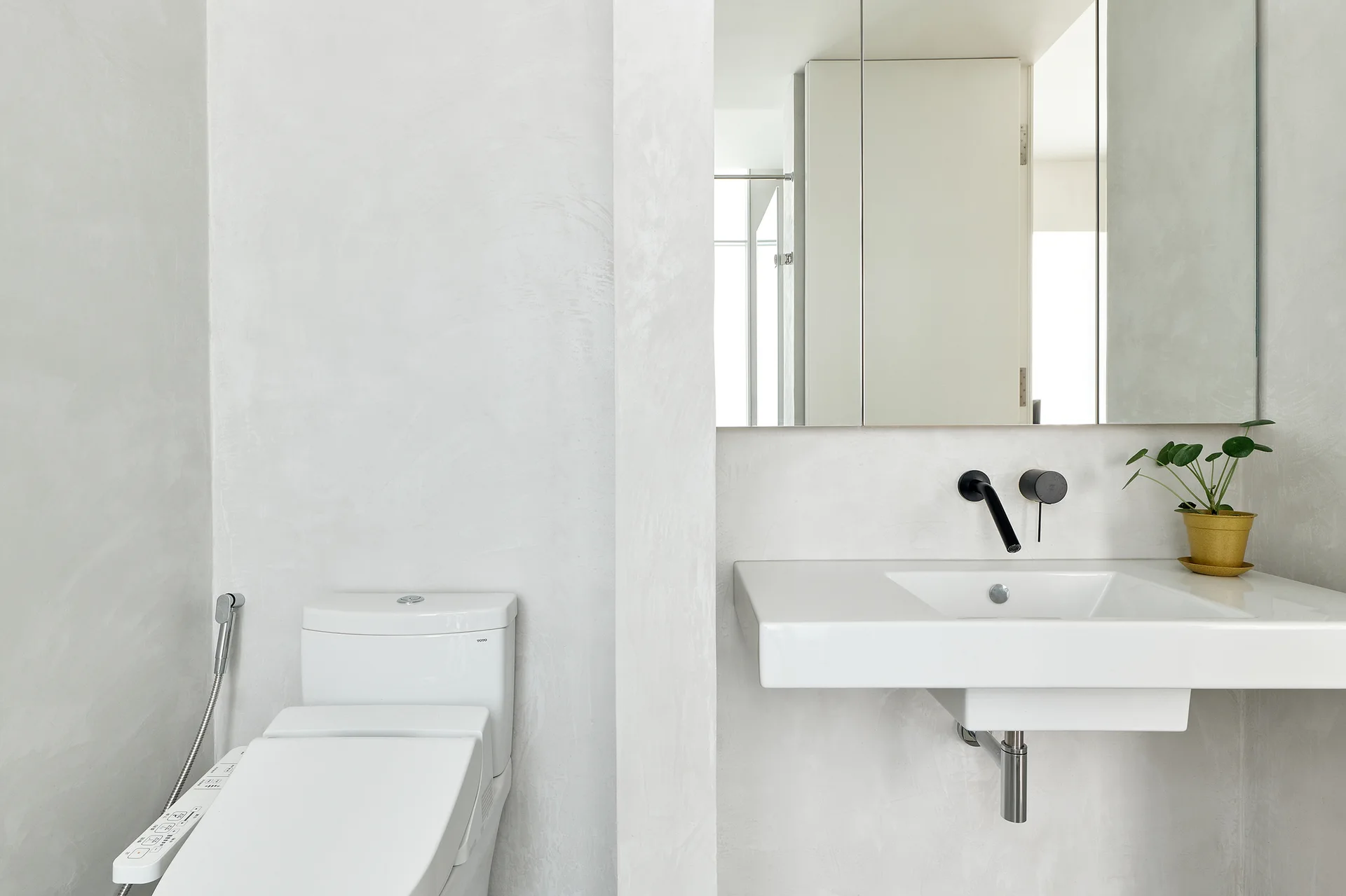 A minimalist bathroom with textured light gray plaster walls, a white wall-mounted sink with a black faucet, and a toilet.