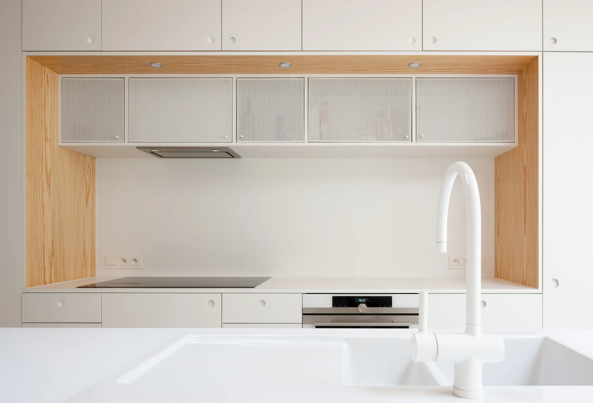 A minimalist kitchen features white handleless cabinets, a natural wood-lined recess with perforated metal doors, and a white arched faucet.