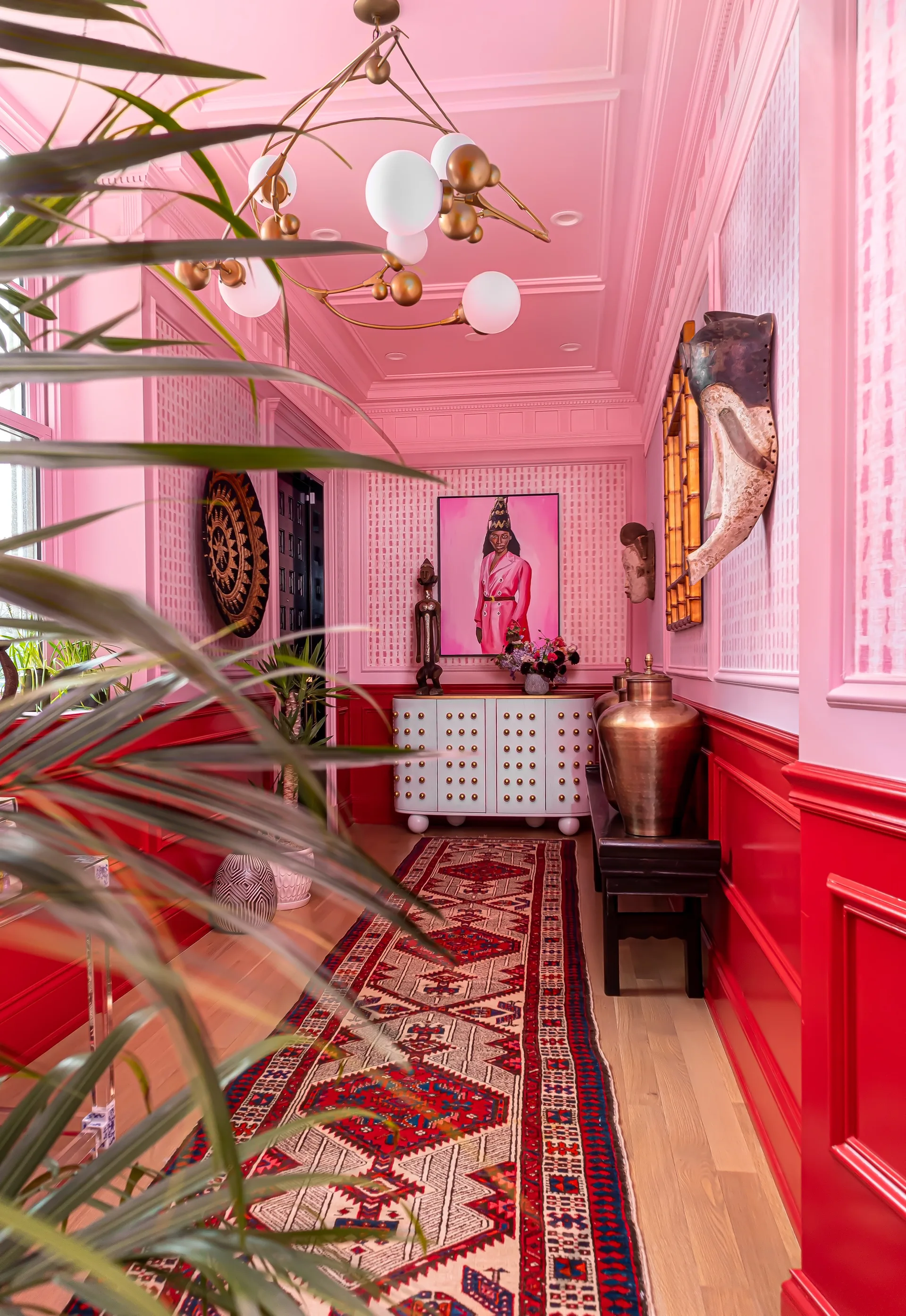 An eclectic hallway features bright pink walls, red wainscoting, a patterned runner rug, a white studded cabinet, and a brass branching chandelier.
