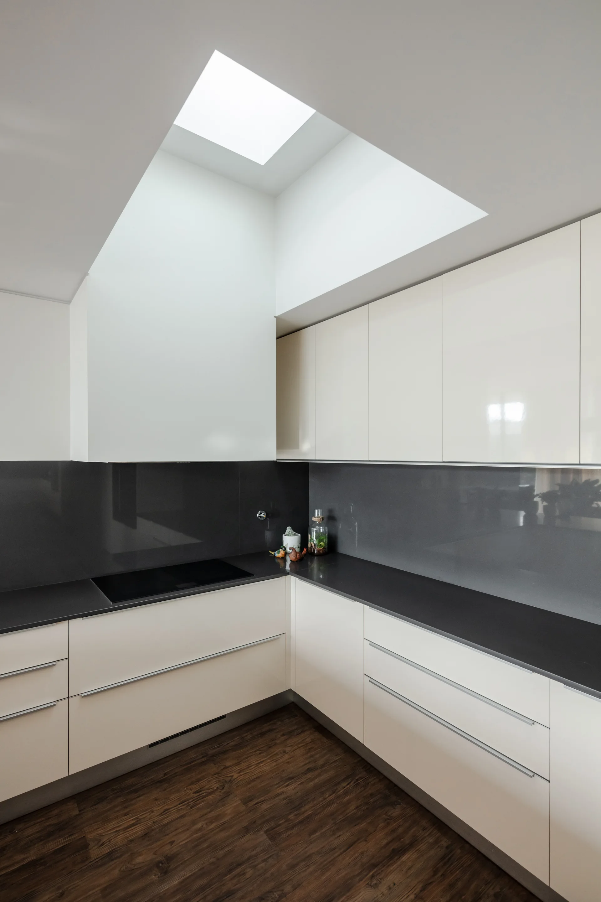 A modern kitchen with glossy cream cabinets, dark charcoal countertops, a reflective grey backsplash, and a bright overhead ceiling skylight.