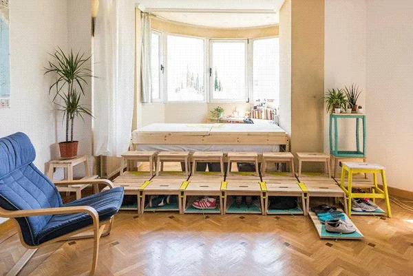A light-filled bedroom features a wooden platform bed by a bay window, a blue armchair, and rows of small wooden stools acting as shoe storage.
