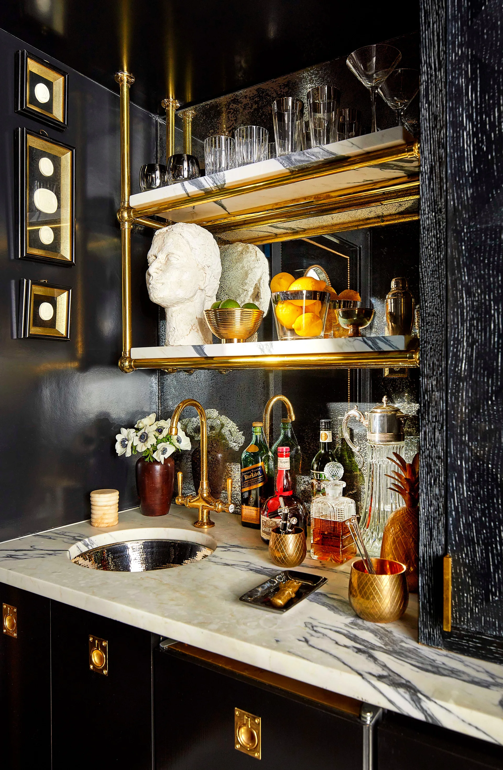 A dark home wet bar features a veined marble countertop, a hammered brass sink, and suspended brass shelves displaying glassware.