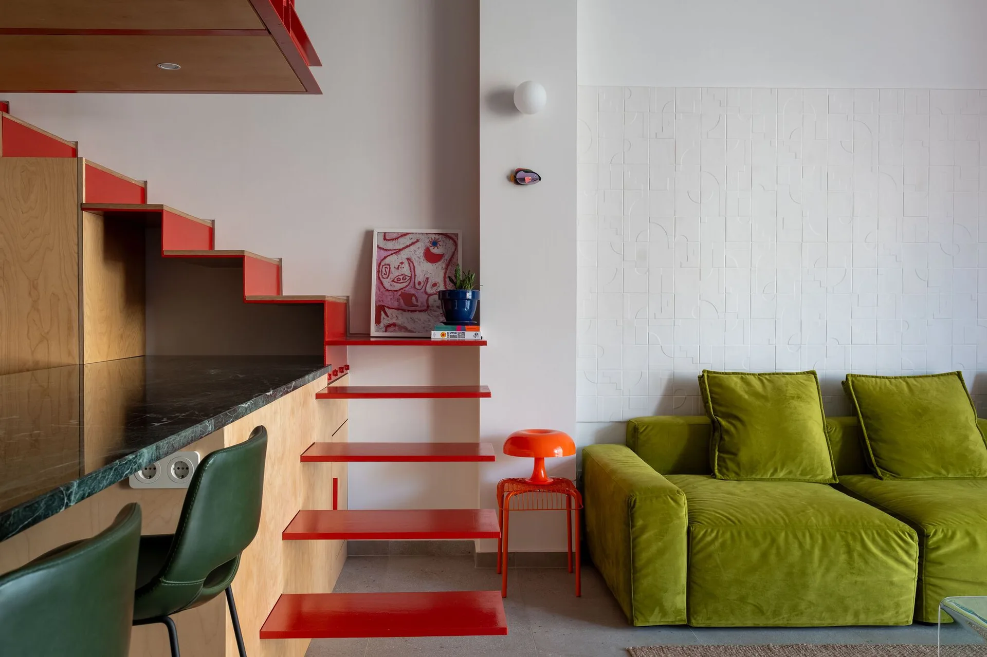 A modern living room features a bright green velvet sofa, a textured white geometric wall, and a custom red and wood staircase with integrated storage.