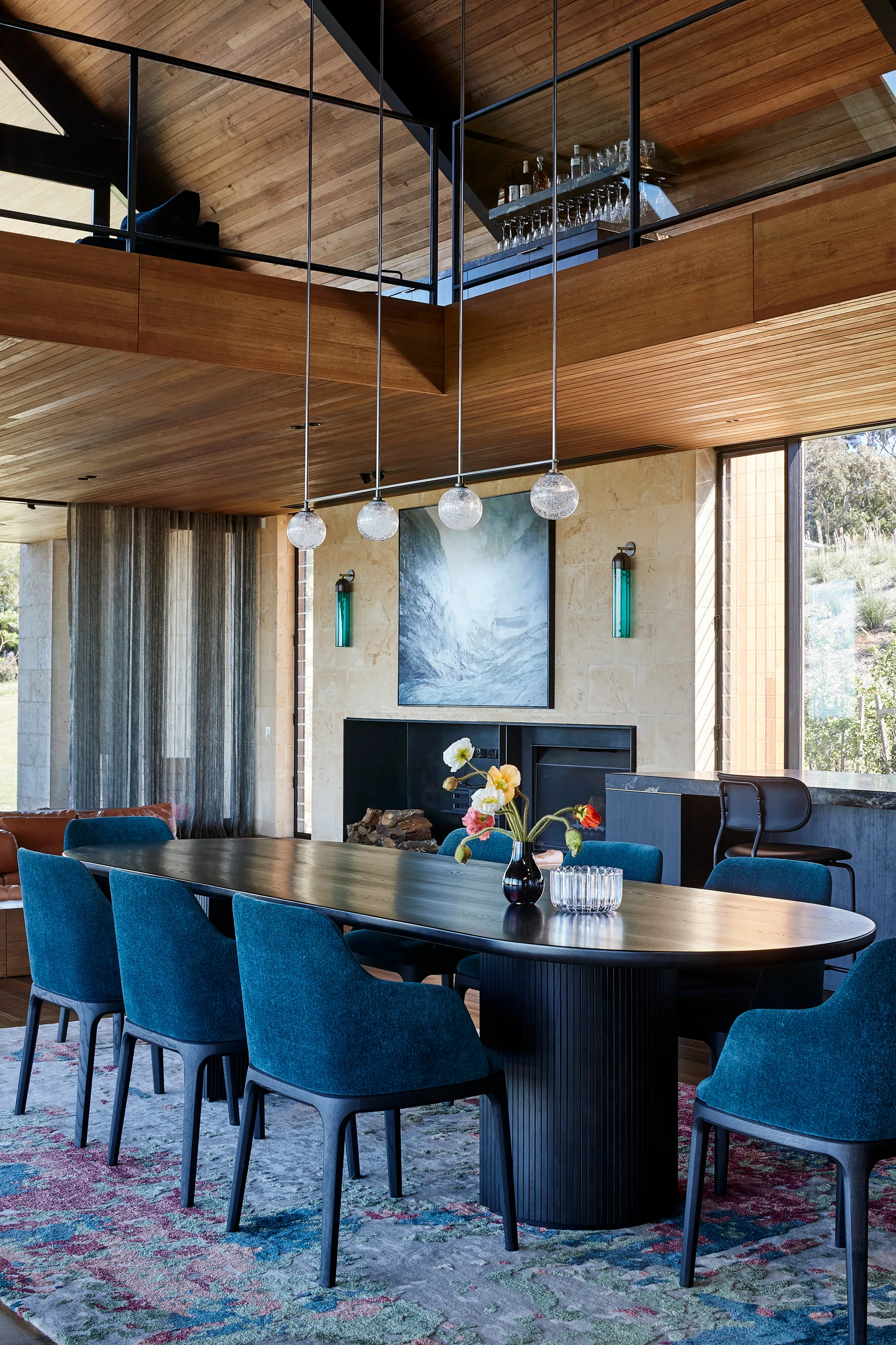 A modern dining room features a dark oval wood table and teal upholstered chairs beneath a warm wood plank ceiling and mezzanine.
