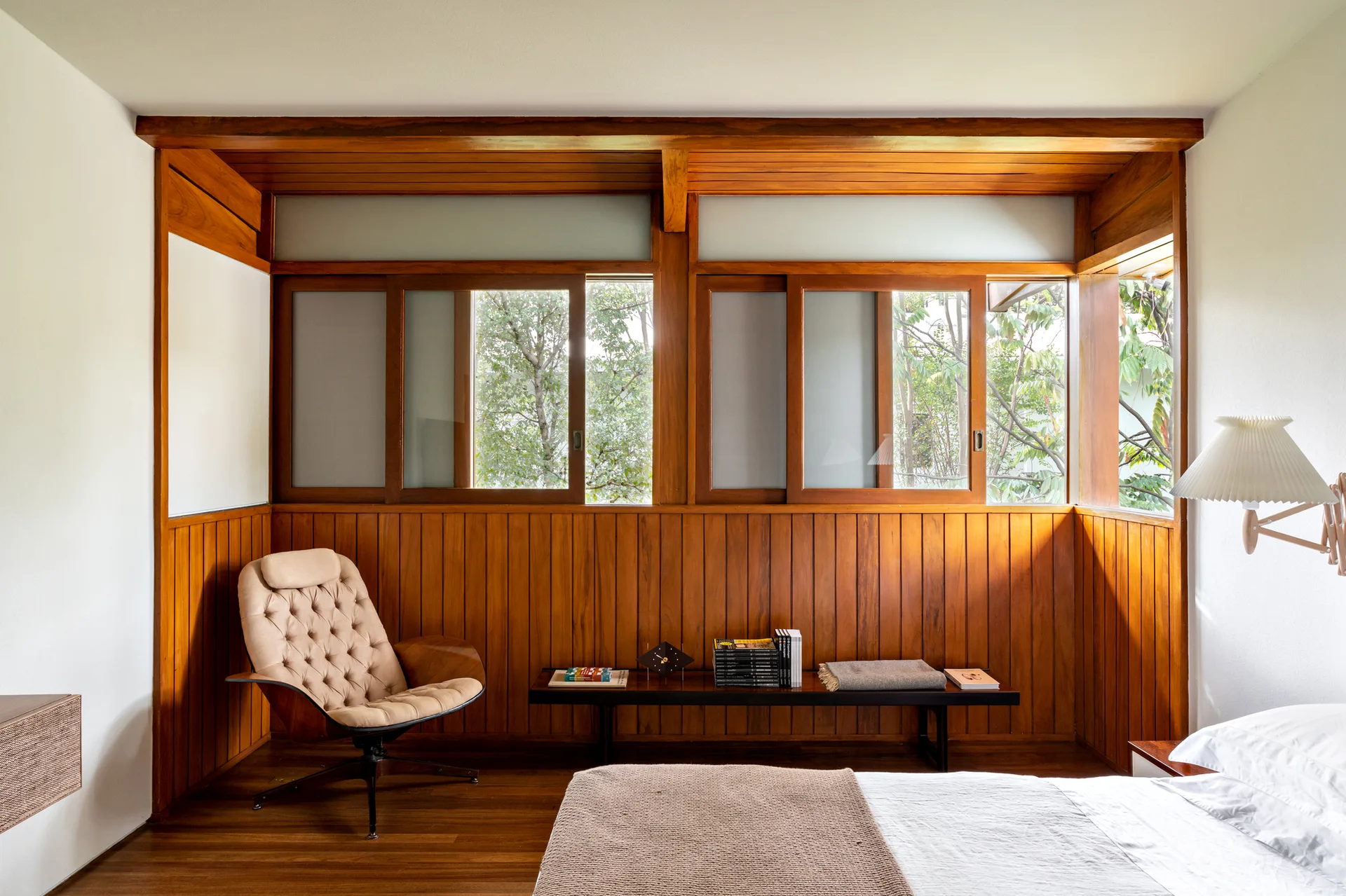 A mid-century bedroom featuring warm vertical wood wall paneling, a tufted lounge chair, and a low wooden bench beneath sliding glass windows.