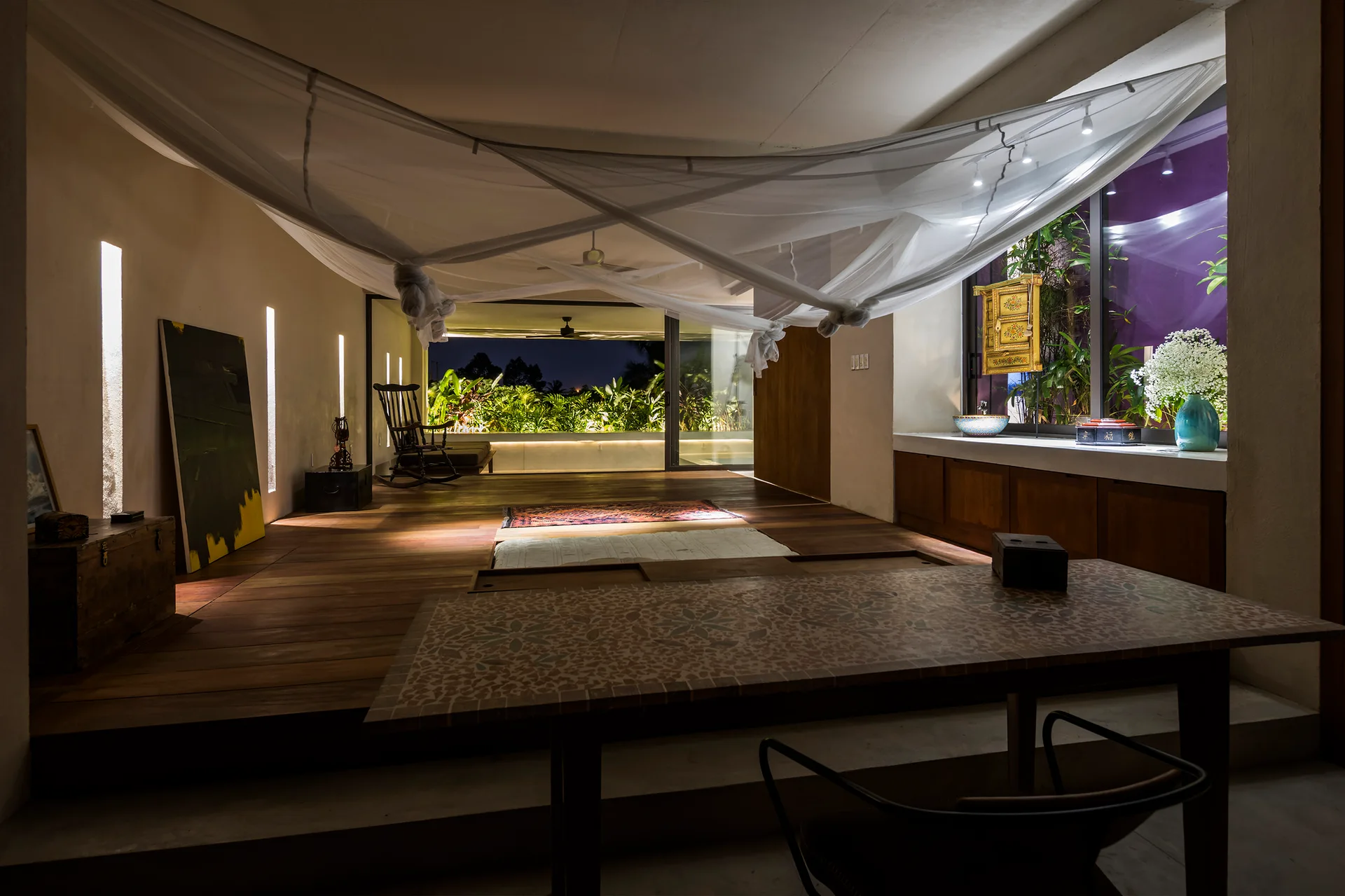 A spacious tropical interior features dark wood floors, a sheer white fabric canopy draped overhead, and a mosaic tabletop in the foreground.
