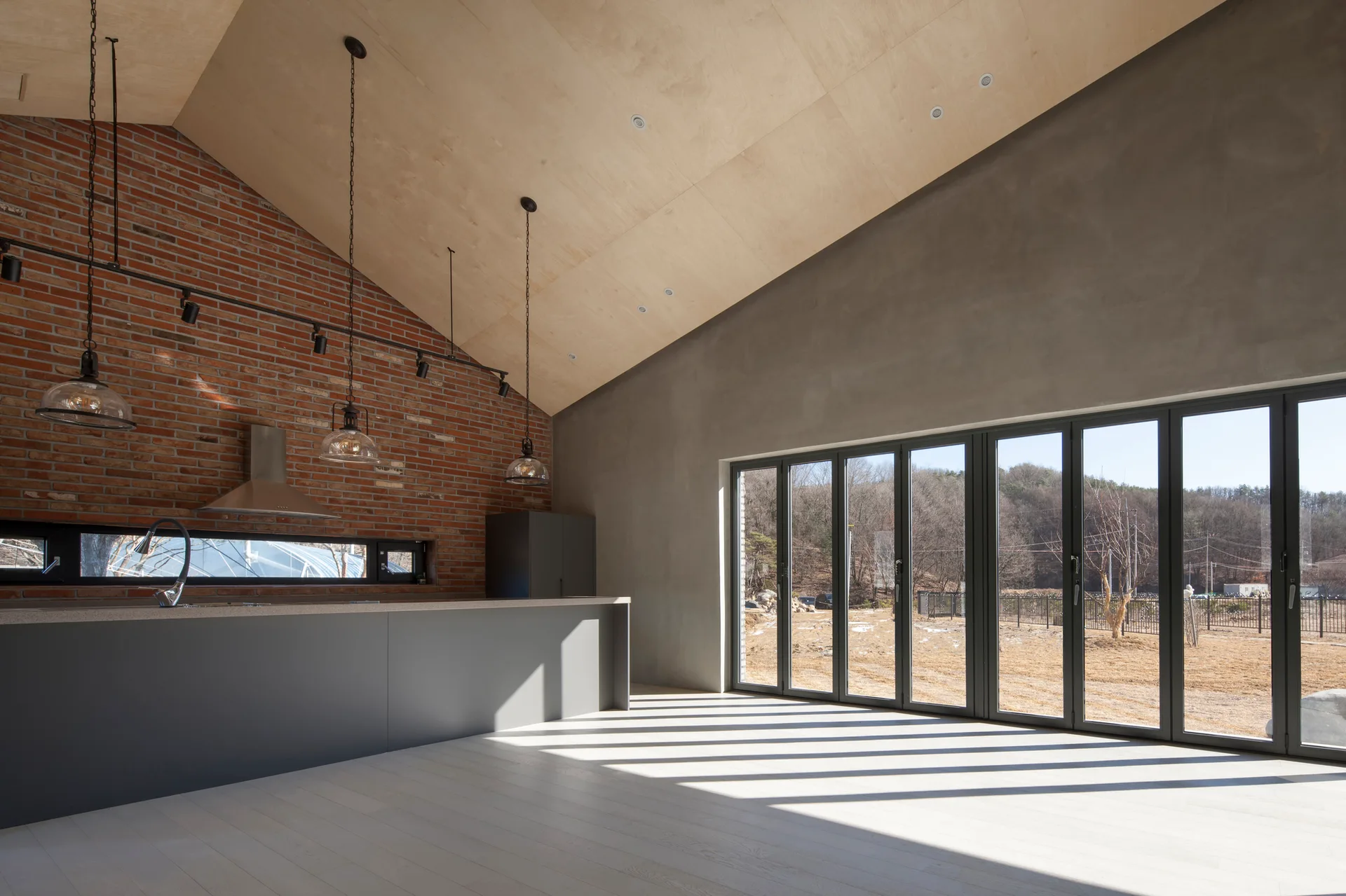 A minimalist industrial kitchen featuring a grey island, an exposed red brick wall, a vaulted plywood ceiling, and black-framed folding doors.