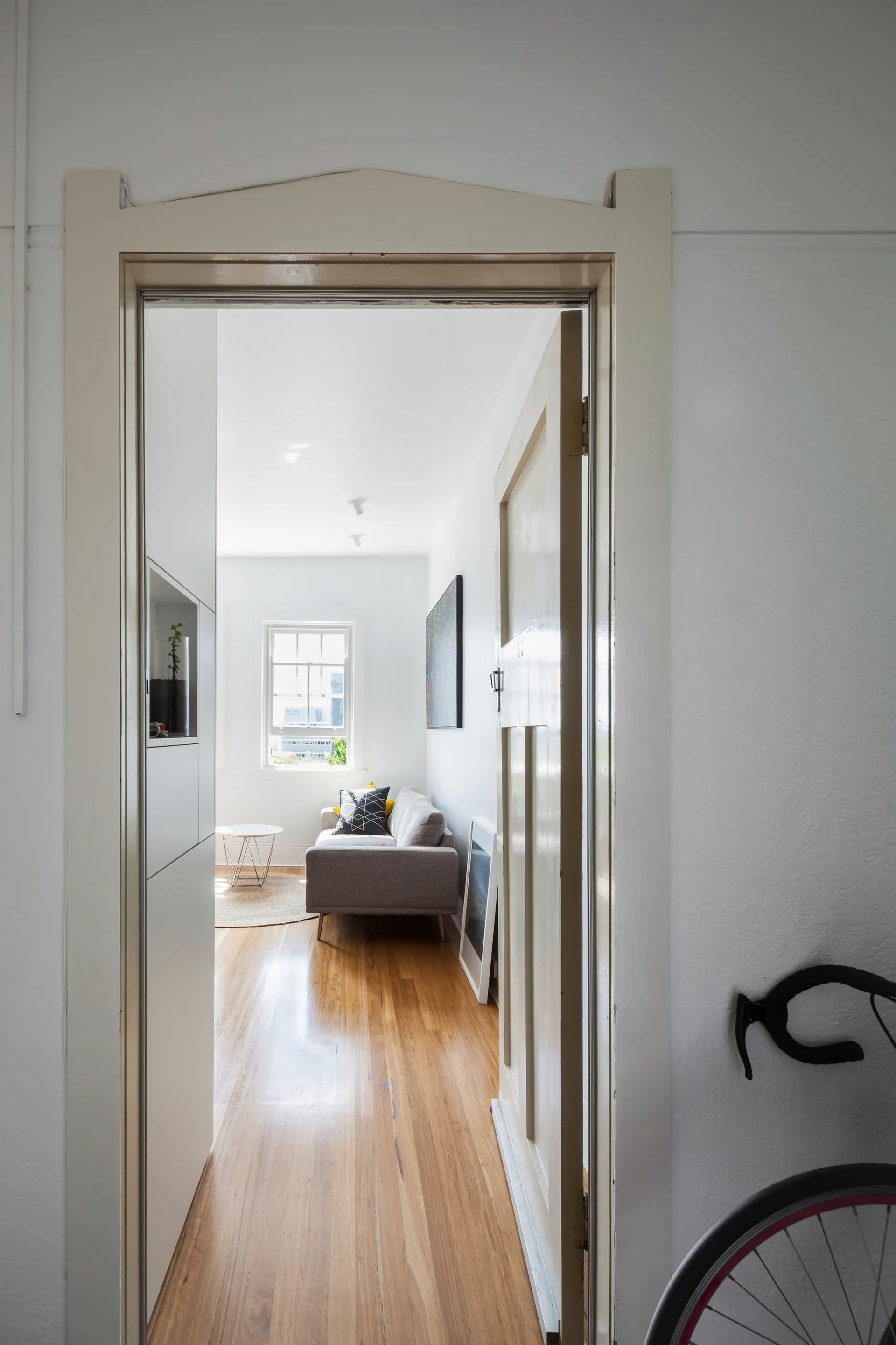 A view through an open doorway into a bright, narrow living room featuring a gray sofa, polished hardwood flooring, and a single far window.