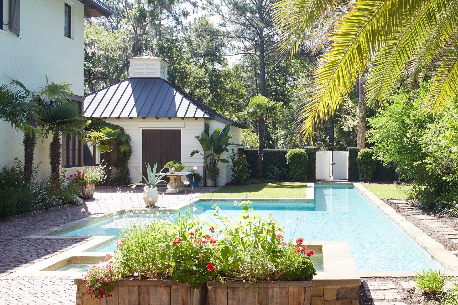 A sunny backyard features a blue L-shaped swimming pool surrounded by a herringbone red brick patio, a white pool house, and lush green palms.