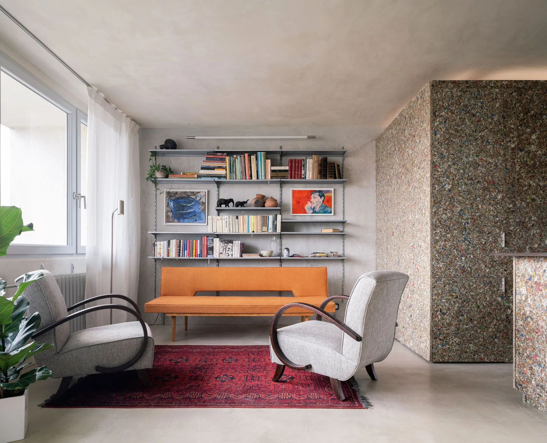 A living room features an orange bench sofa, grey curved armchairs, a patterned red rug, and a textured, multi-colored partition wall.