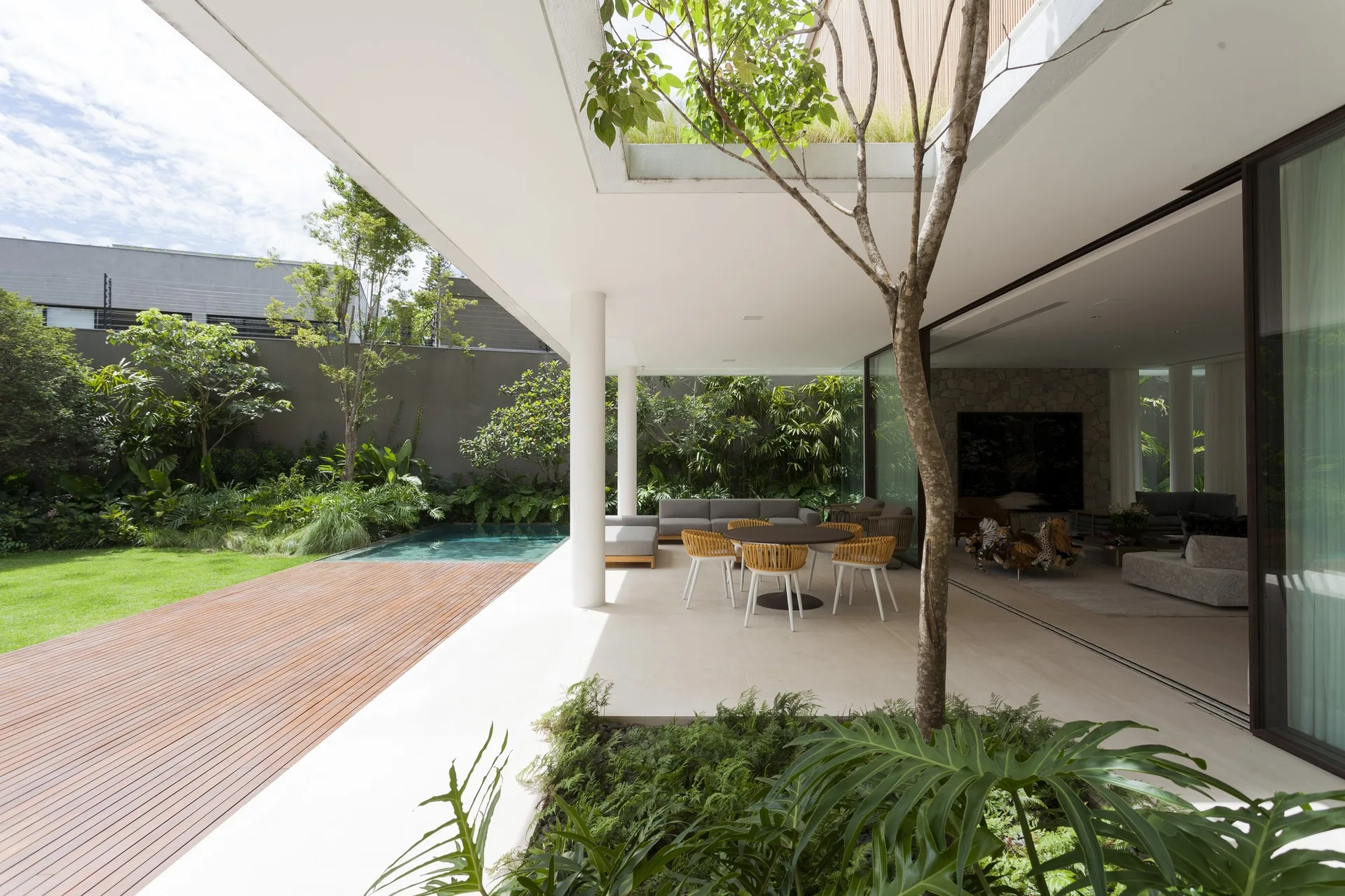 A modern covered patio with a dining set and lounge furniture sits next to a swimming pool, wood deck, and lush garden.