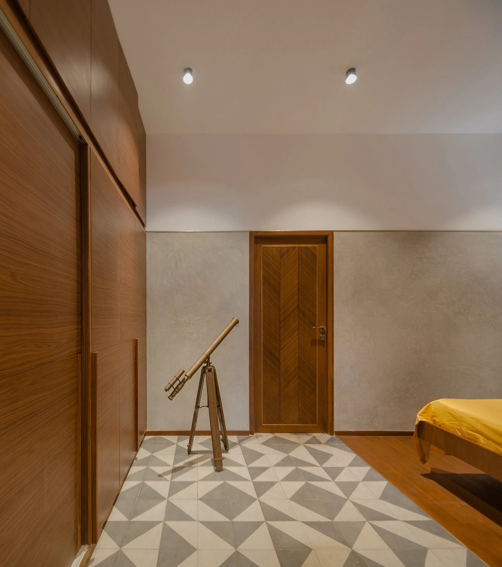 A modern bedroom features a large wood veneer wardrobe, a matching herringbone pattern wood door, and a grey and white geometric tiled floor.