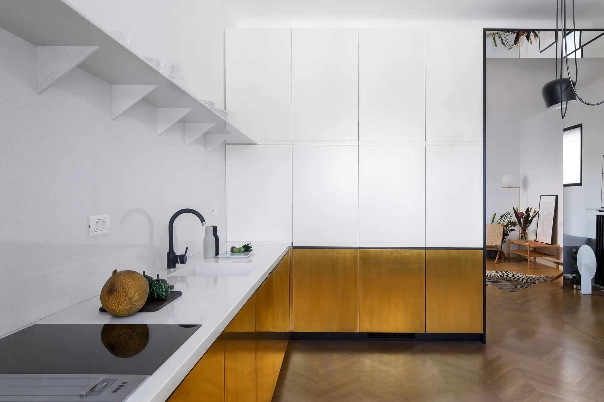 A minimalist kitchen featuring matte white upper cabinets, reflective gold lower cabinetry, a white countertop, and herringbone wood flooring.