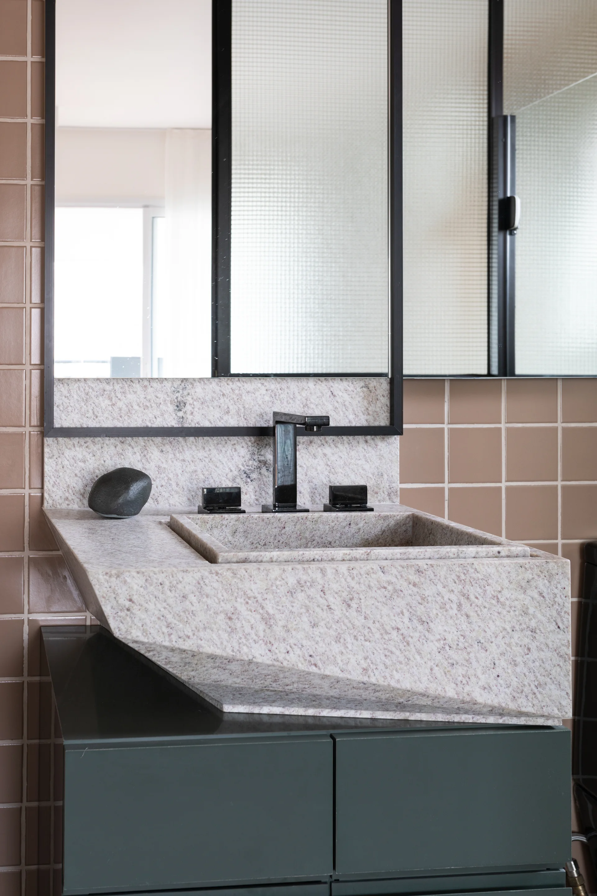 An angular speckled stone bathroom sink rests on a dark green cabinet against a grid of square terracotta wall tiles.