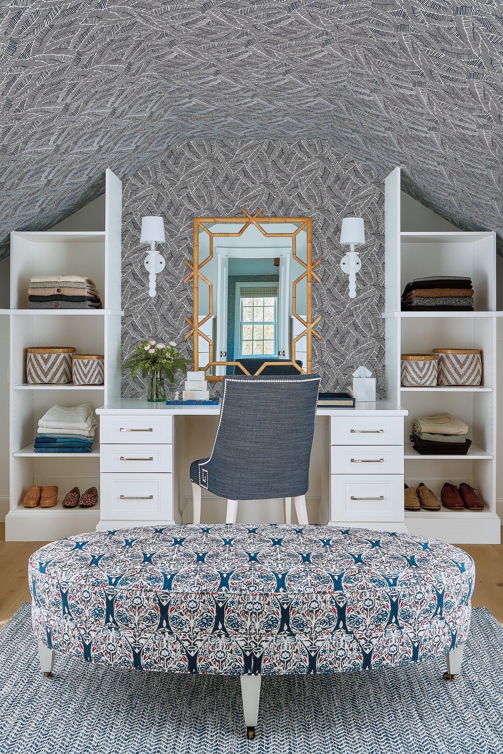 A dressing room in an attic features a white vanity desk flanked by open shelving, under a sloped ceiling covered in blue and white leaf wallpaper.