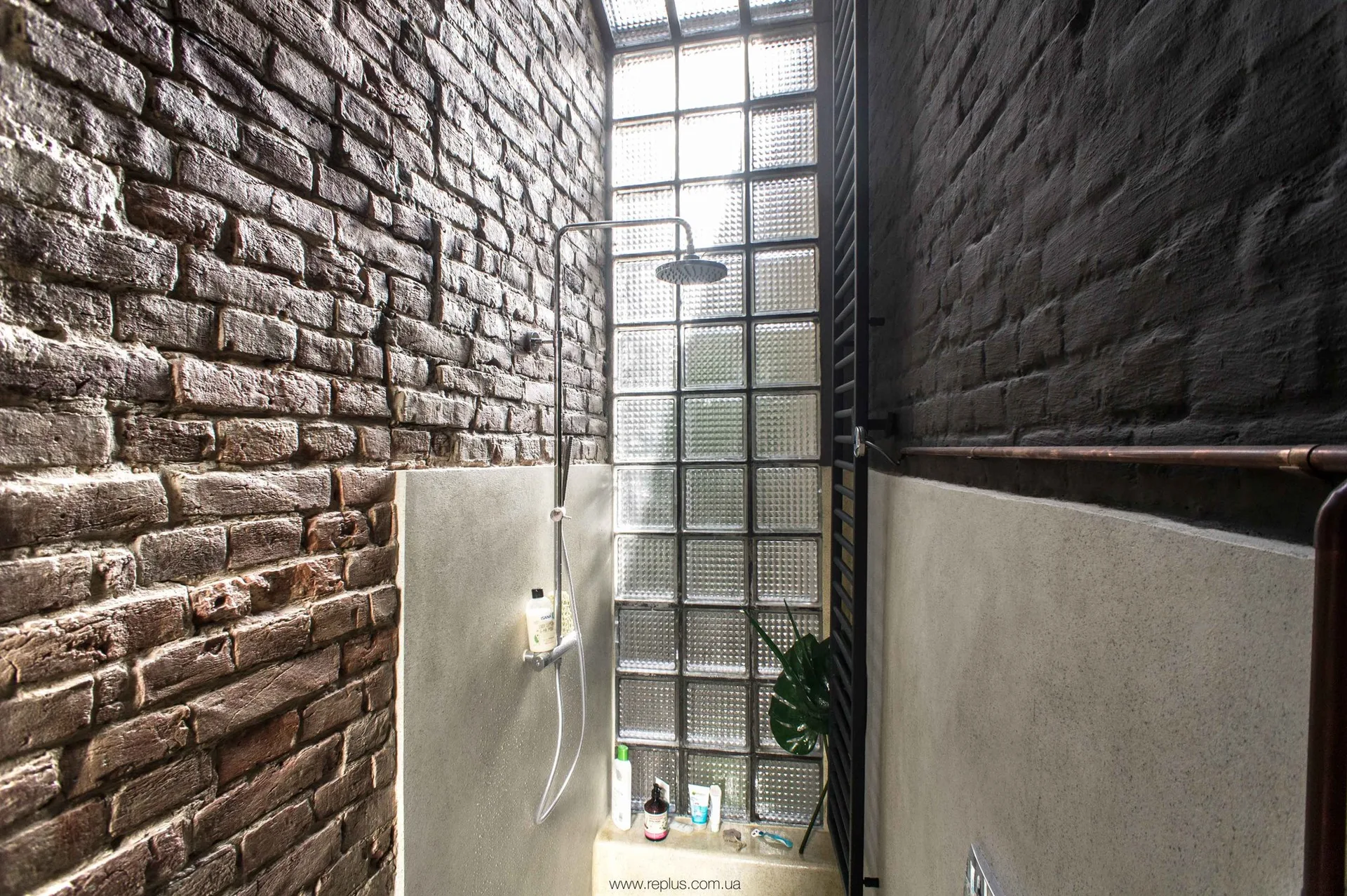 A narrow bathroom shower with dark exposed brick walls, a tall translucent glass block window, and a sleek metal shower fixture.