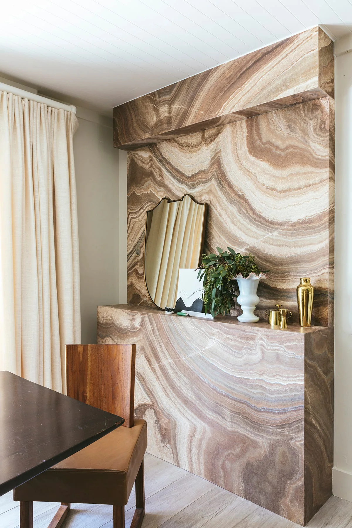 A dining room features a swirling brown and tan stone fireplace surround decorated with a gold shield-shaped mirror, a white vase, and a wooden chair.
