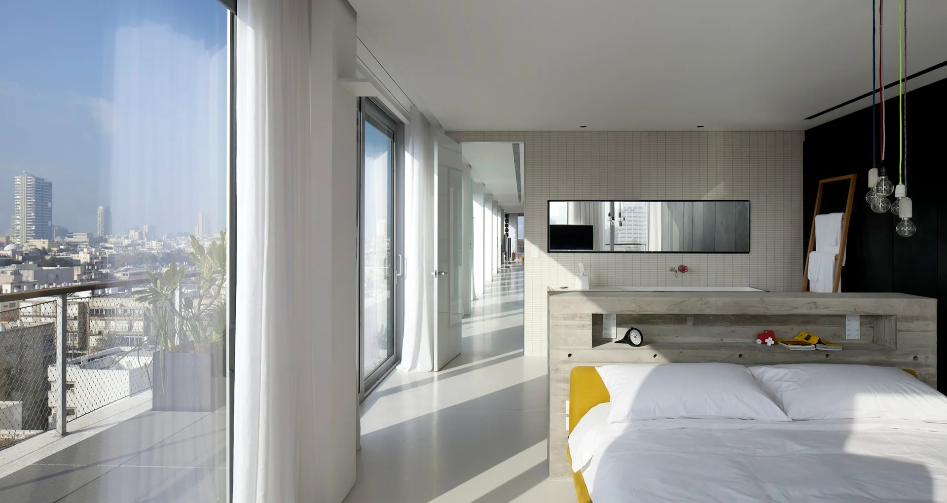 A minimalist bedroom features a low bed with a yellow base and a thick concrete headboard with a built-in shelf next to large glass doors.