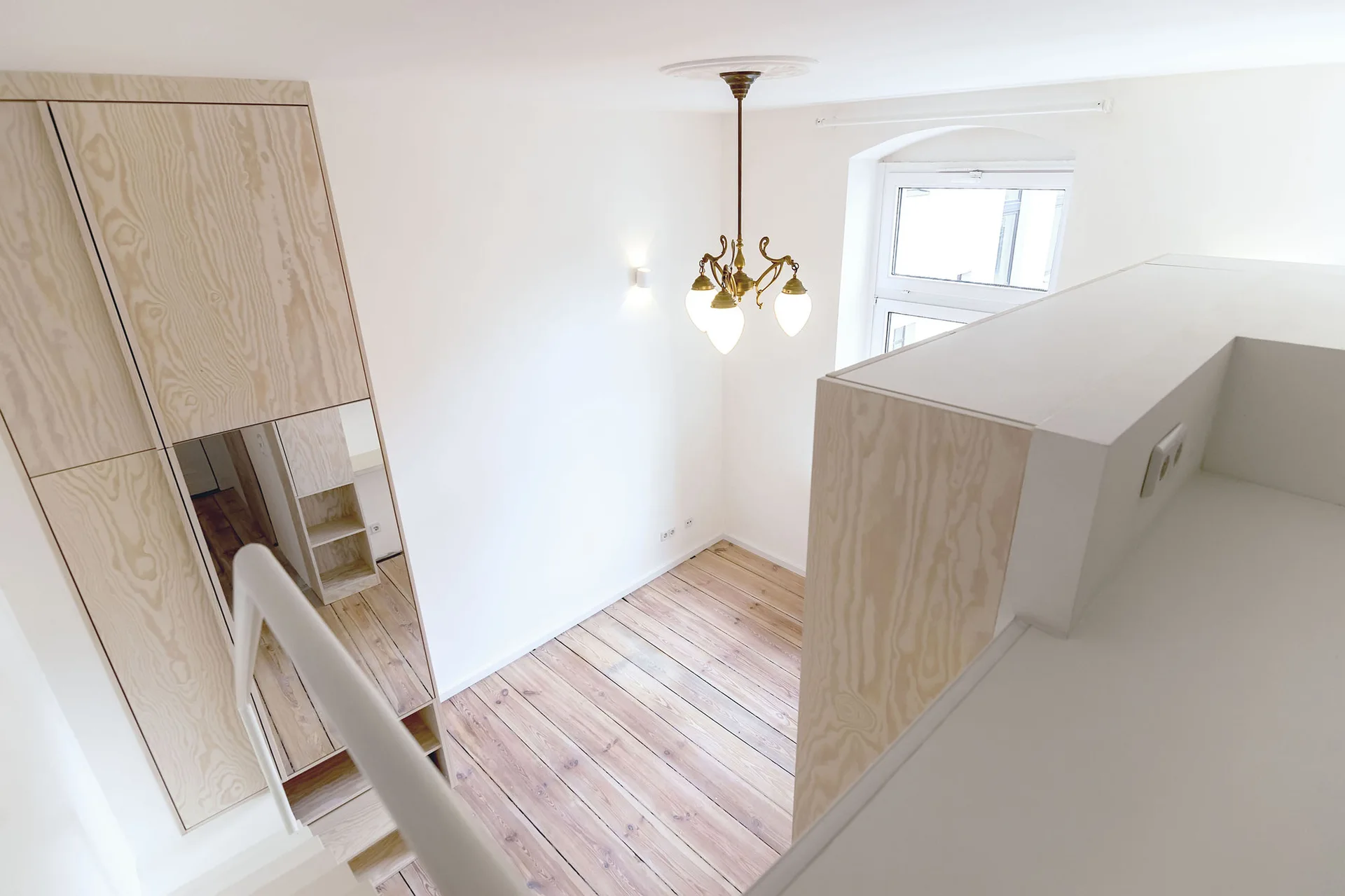 An elevated view looking down into a white minimalist room with light wood built-in cabinetry, plank wood floors, and a brass chandelier.