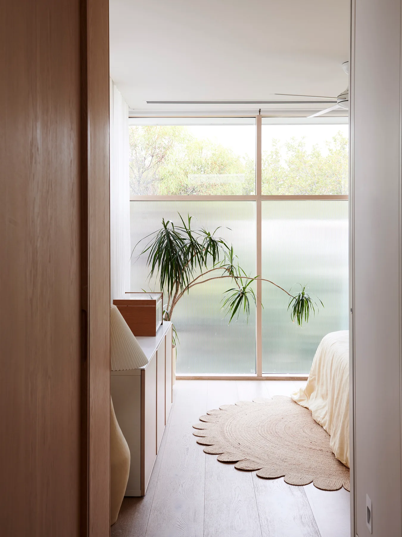 A light-filled bedroom viewed through a doorway features a large fluted glass window, a scalloped jute rug, a houseplant, and a low console cabinet.