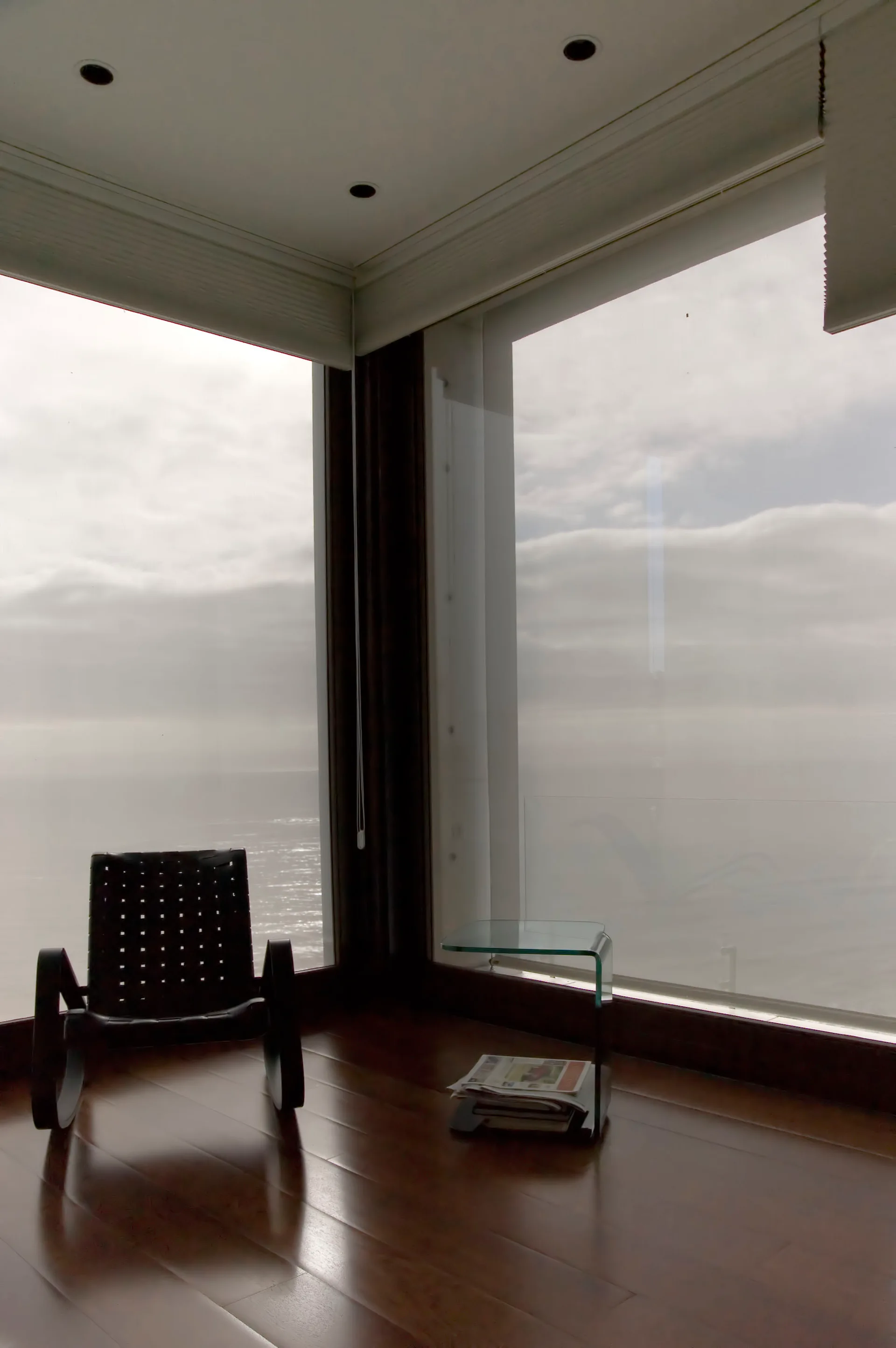 A black woven rocking chair and a curved glass side table rest on polished dark wood floors next to large corner windows overlooking the ocean.