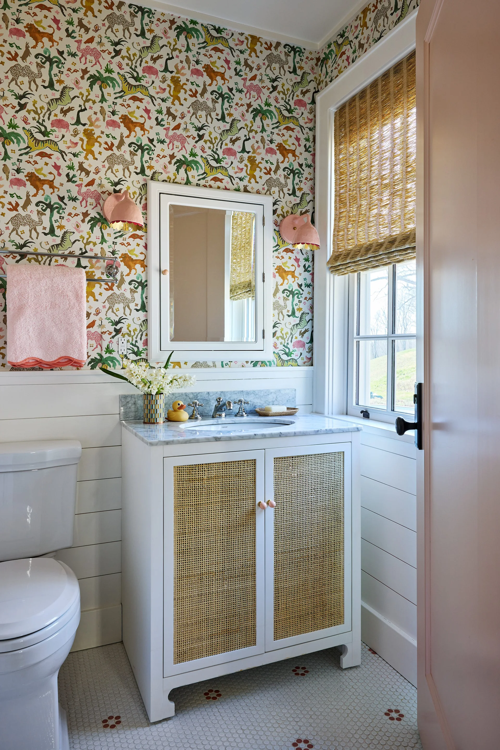 A bathroom featuring a white vanity with cane door panels, colorful animal-themed wallpaper, white wood wainscoting, and a window with a woven shade.