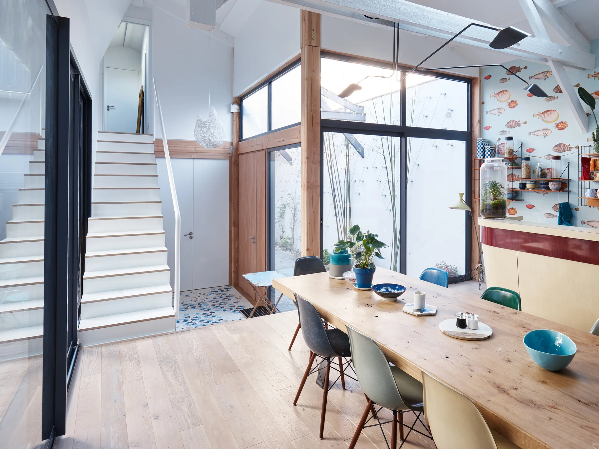 A bright dining room features a long wooden table surrounded by colorful chairs, next to a white staircase and large wood-framed windows.