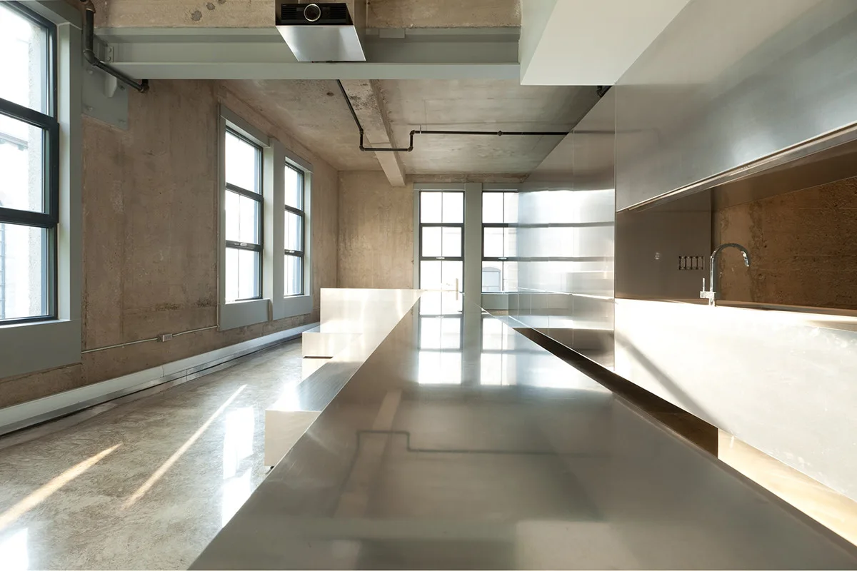 An industrial loft kitchen featuring a long stainless steel island, matching metal cabinetry, raw concrete walls, and large grid windows.