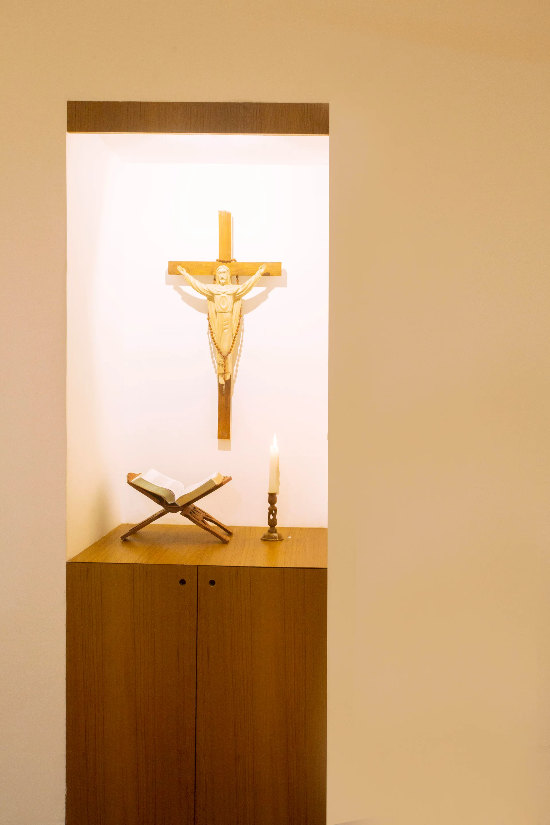 A wooden cabinet is built into a beige recessed wall niche featuring a crucifix, an open book on a wooden stand, and a single lit candle.