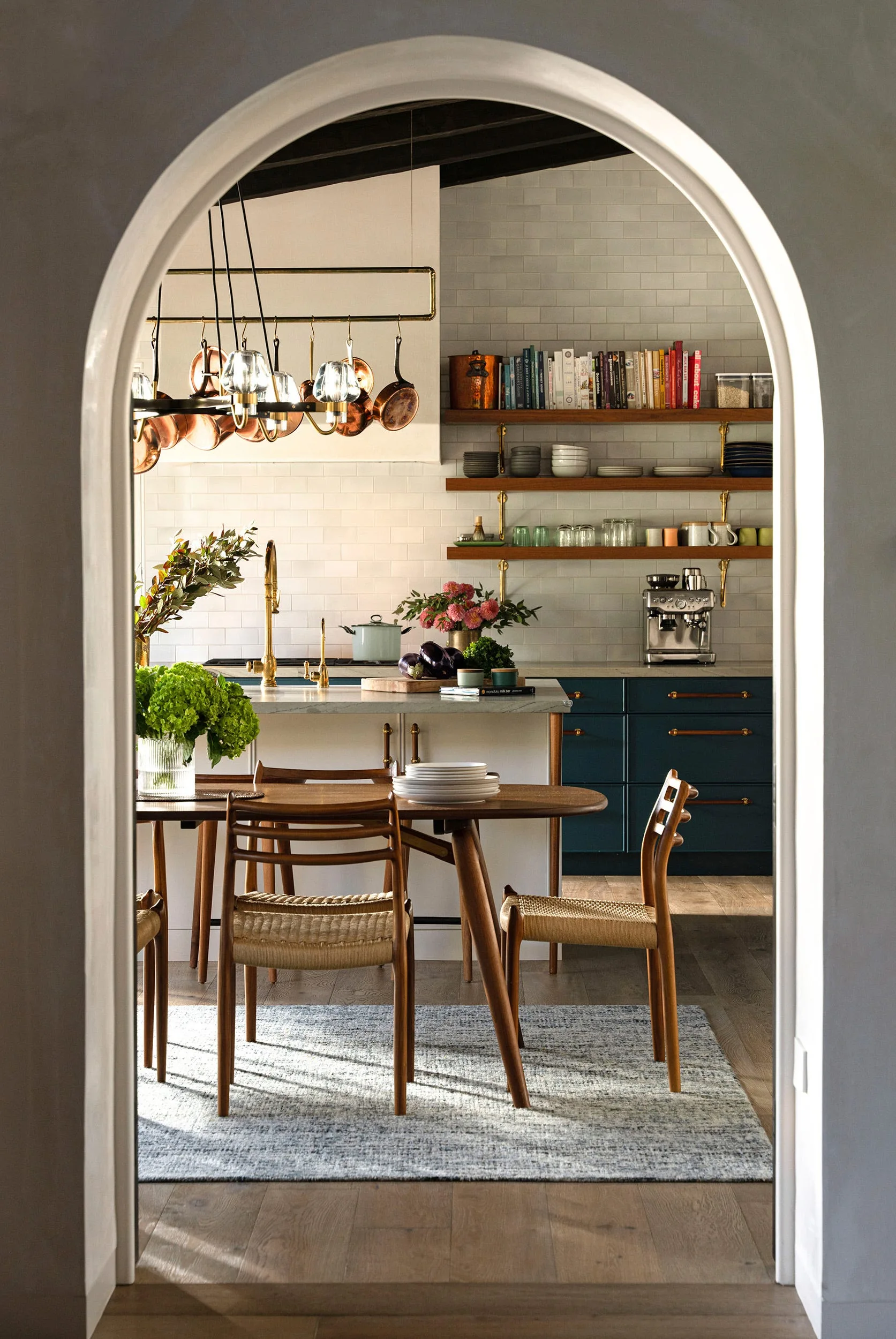 A white archway frames a view of a dining table on a blue rug in front of a kitchen with navy cabinetry and open wood shelves.