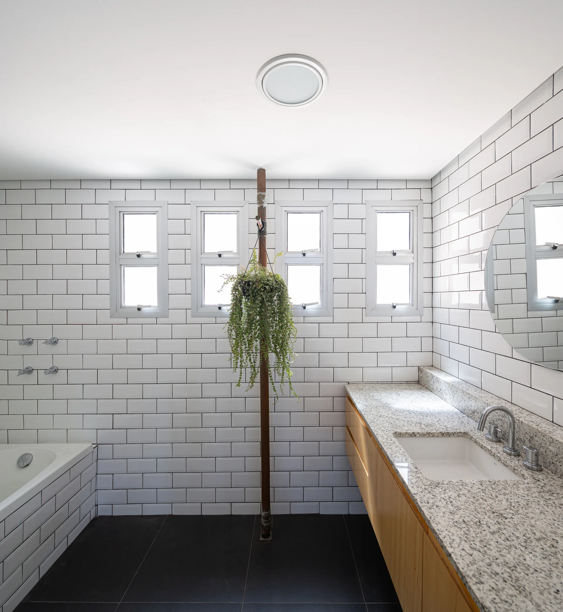 A modern bathroom features white subway tile walls, a wood vanity with a speckled countertop, a circular mirror, and a hanging plant on a central pipe.
