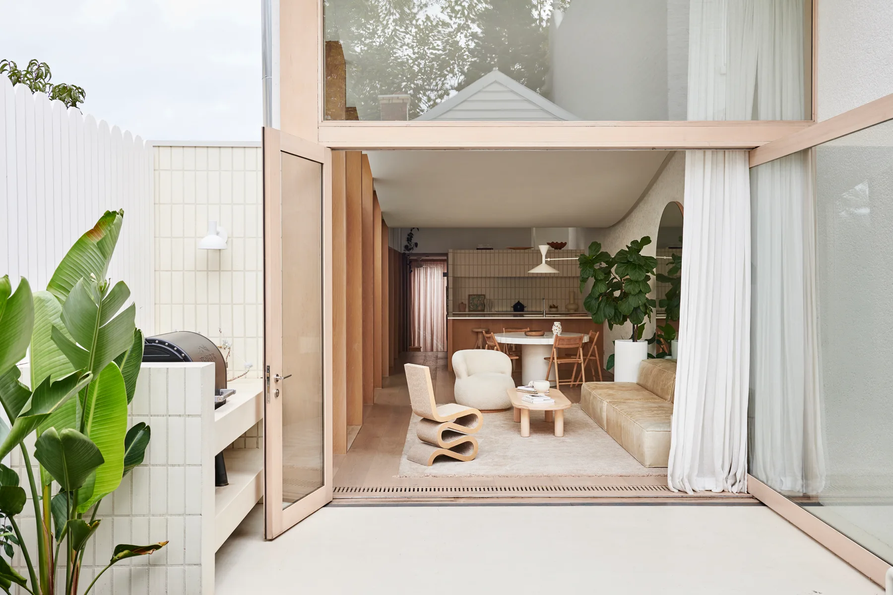 A bright open plan living space with light wood floors and low-profile neutral furniture opening up to an outdoor patio through massive glass doors.